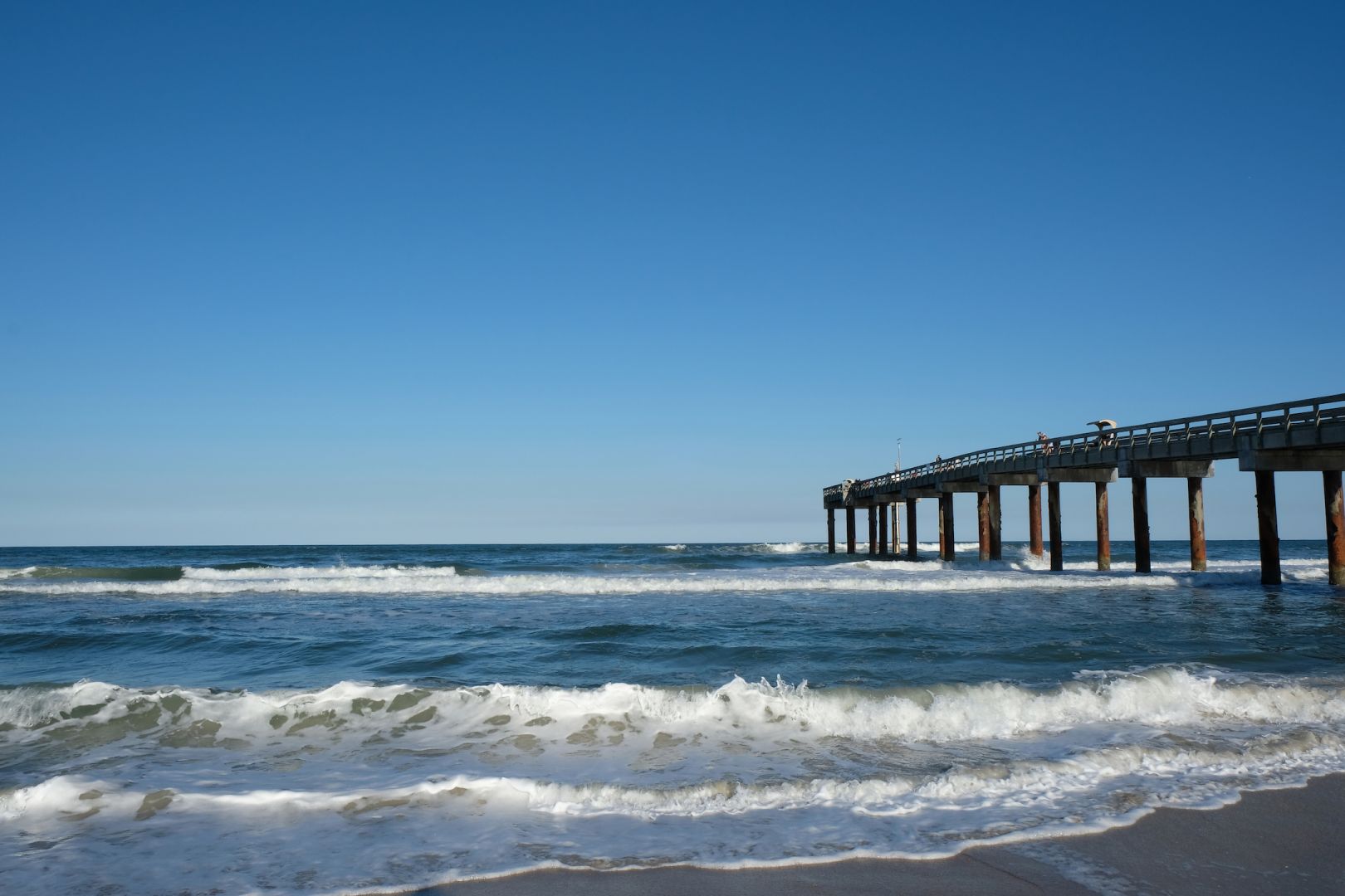 Ocean waves gently crashing against a long pier under a clear blue sky.