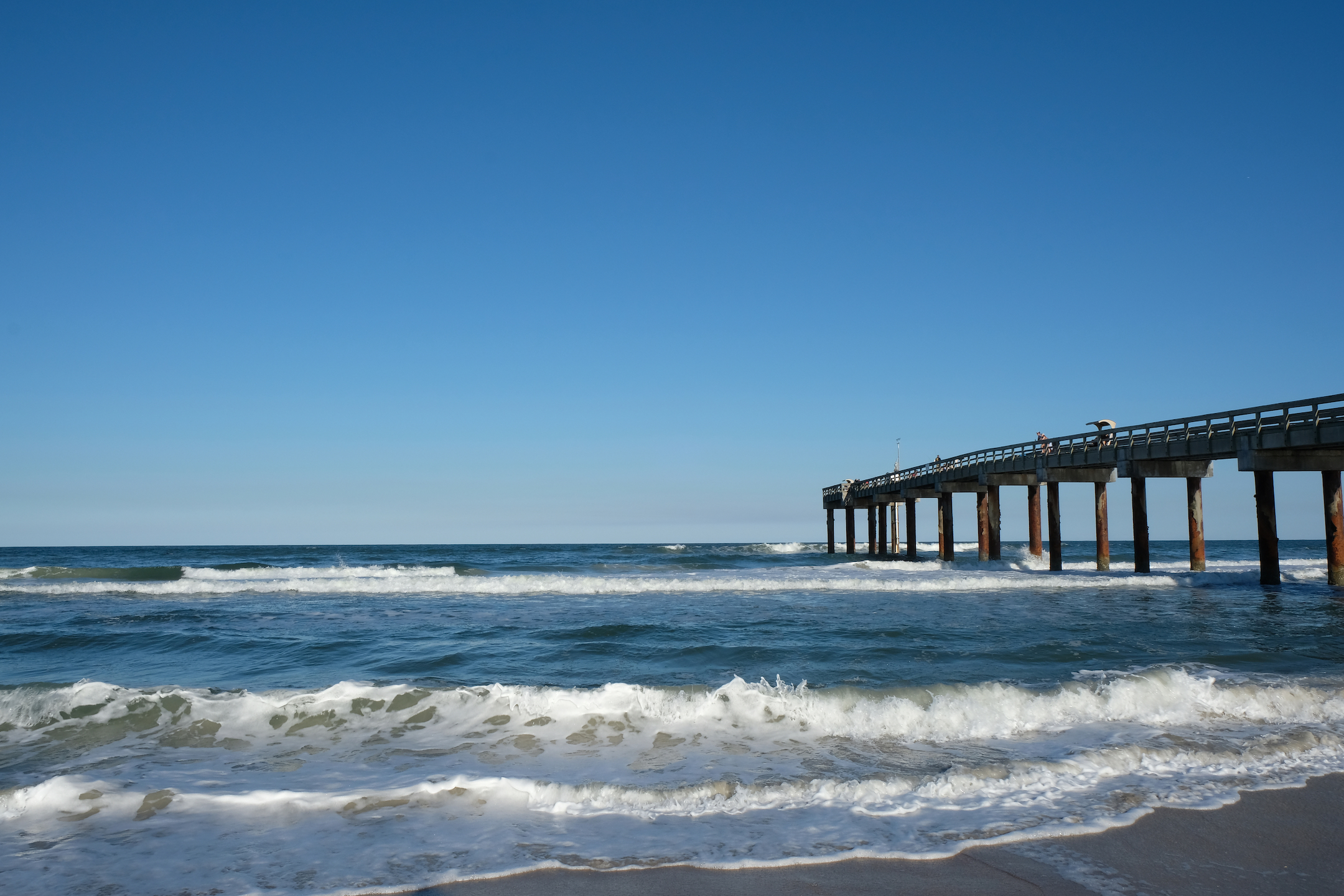Ocean waves gently crash against a sandy beach with a long pier extending into the blue sea under a clear sky.