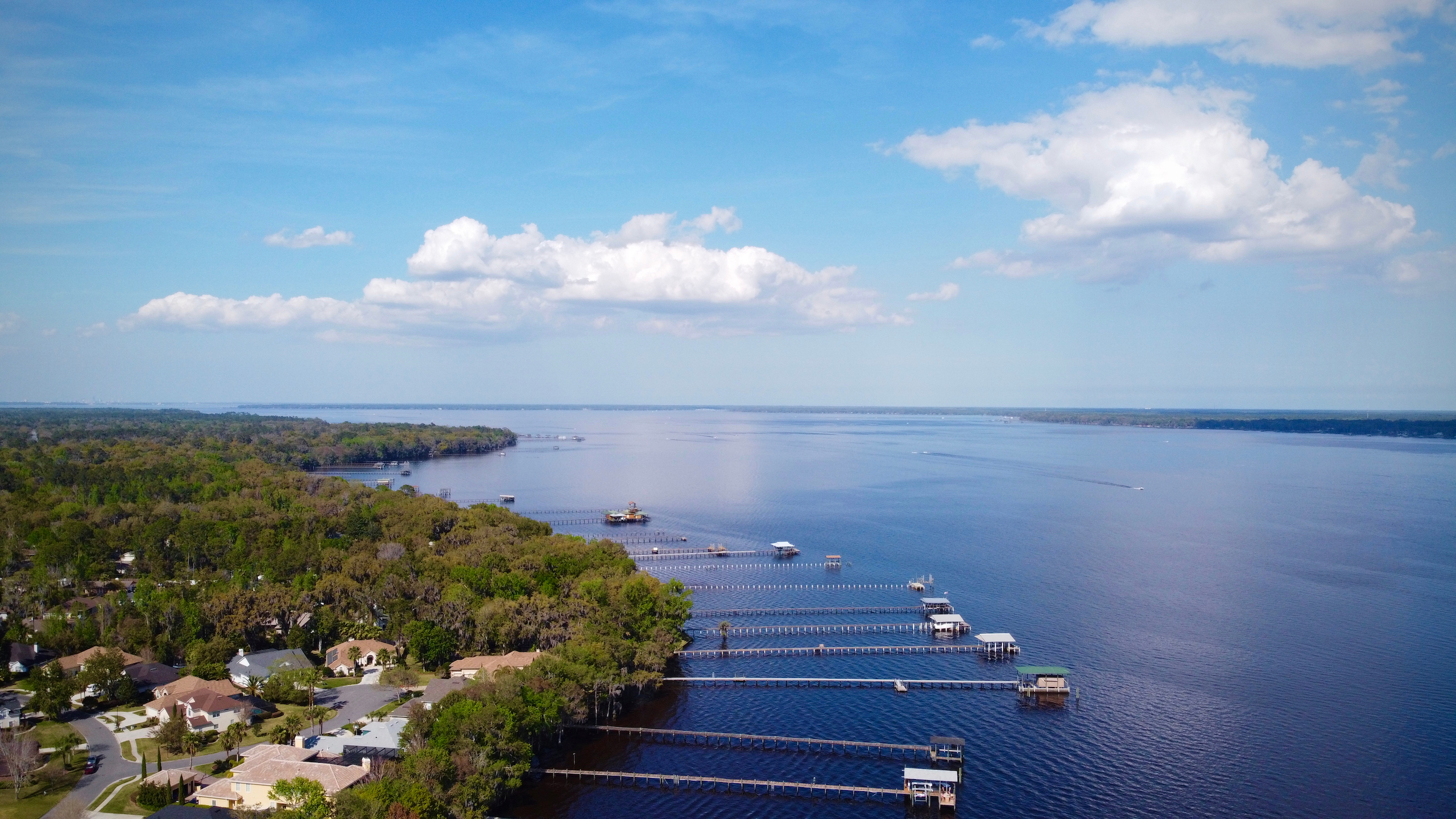 Aerial view of a serene lakefront residential area with docks extending into the calm blue water under a partly cloudy sky.