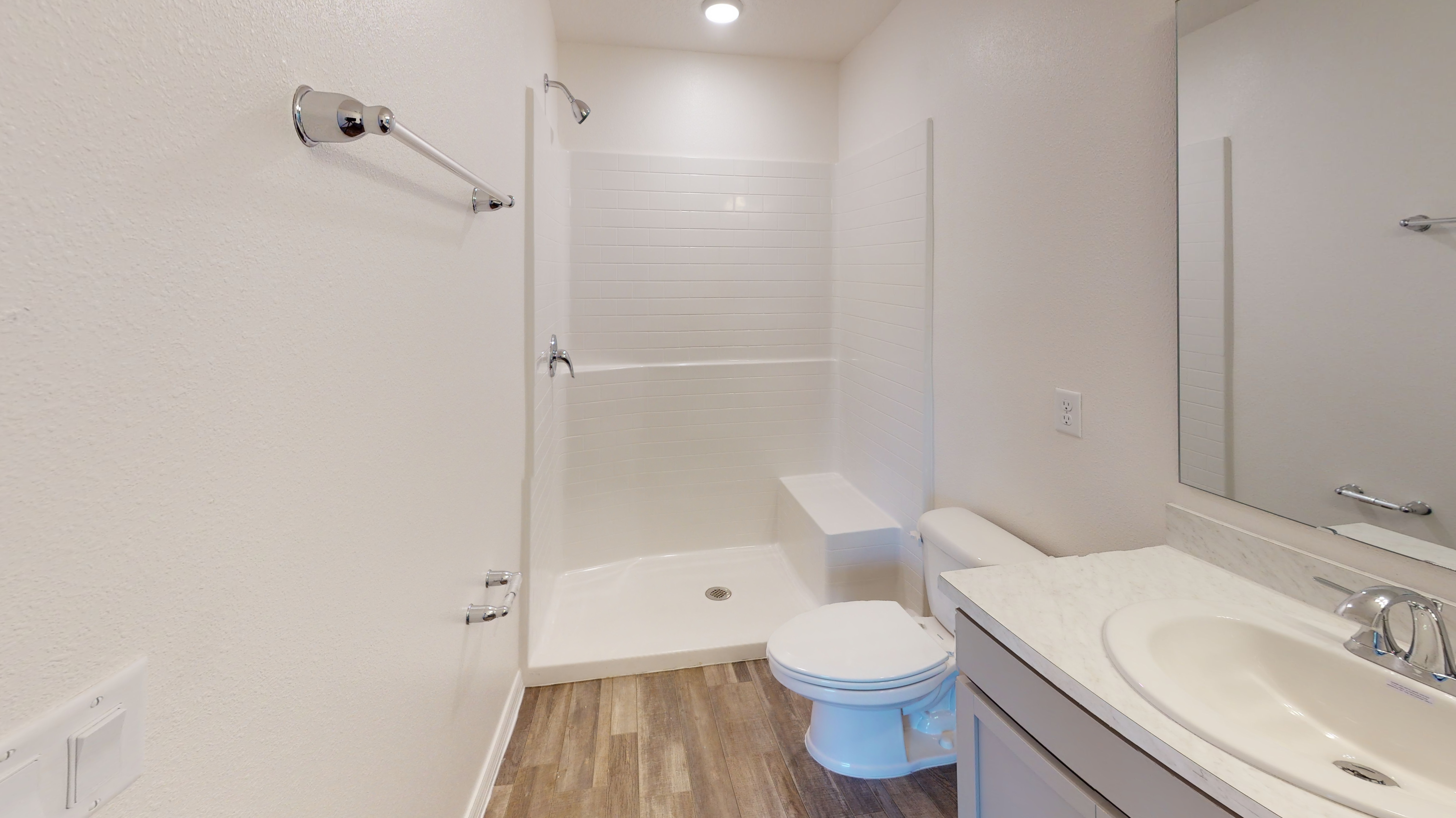 "Modern bathroom with white tiled walk-in shower, toilet, and marble countertop sink on a wood-patterned floor."