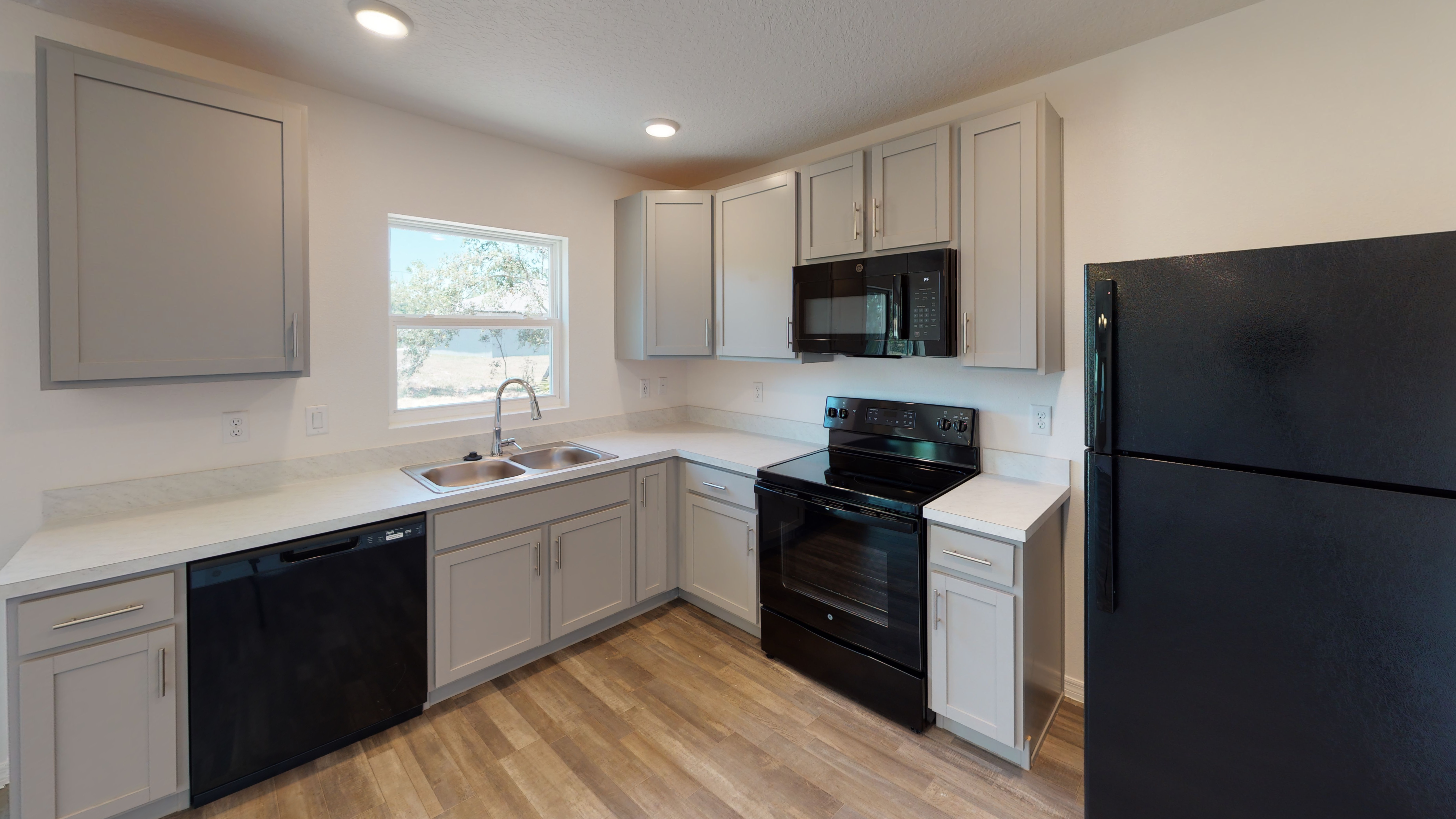 Modern kitchen with gray cabinets, black appliances, and white countertops, featuring a large window and wood-style flooring.