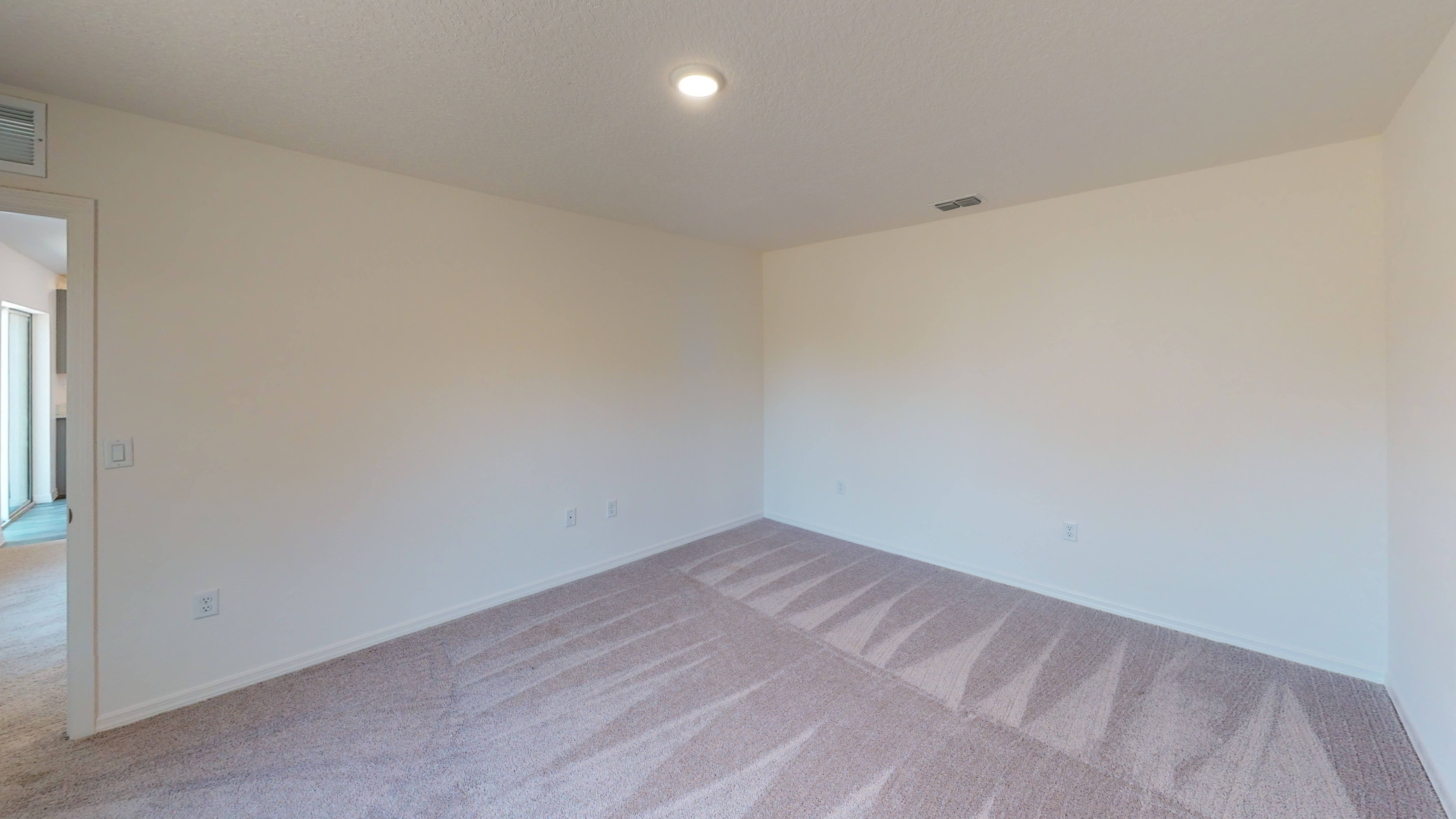 Empty beige-carpeted room with white walls, ceiling light, and adjacent open door leading to a hallway.