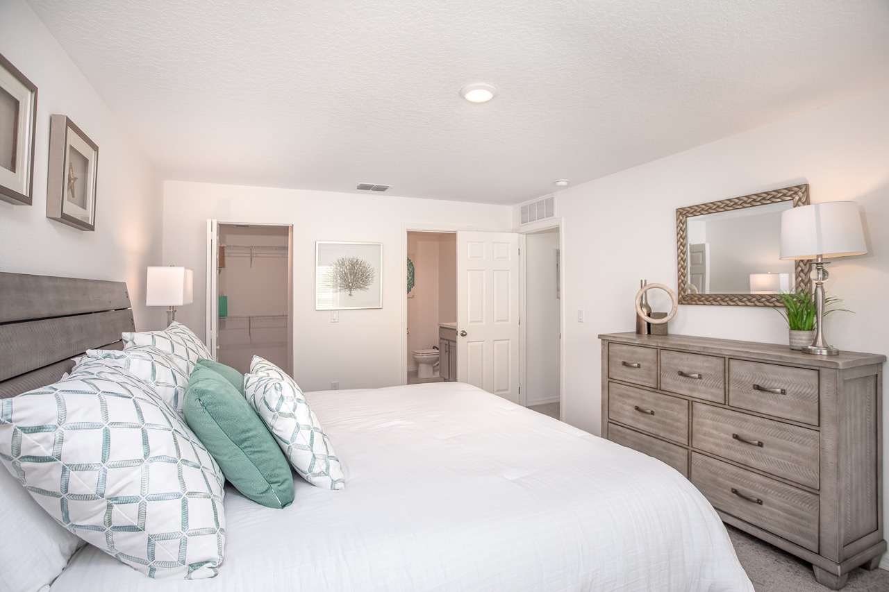 Modern bedroom with a neatly made bed, decorative pillows, wooden dresser, wall art, and a walk-in closet.