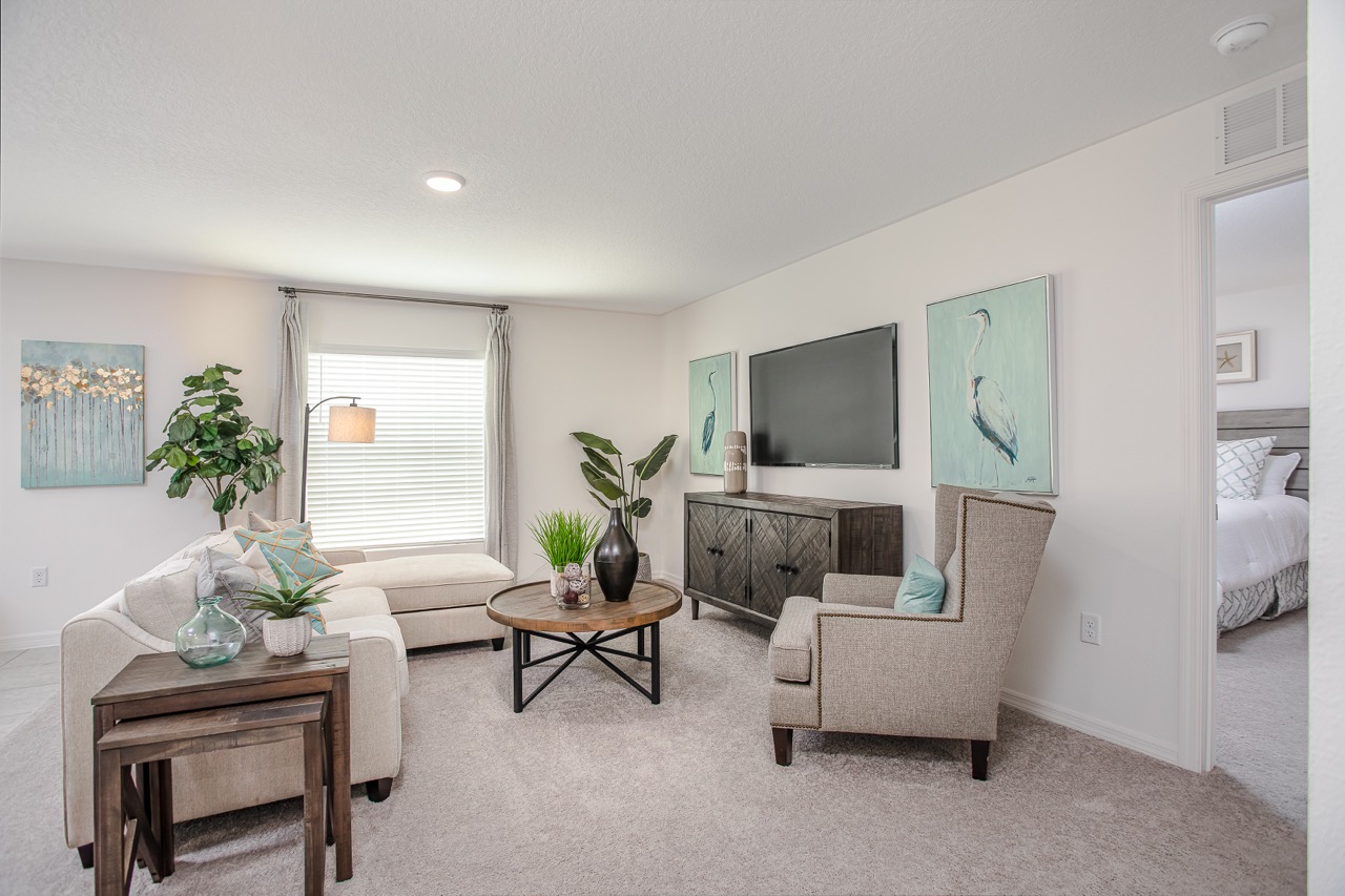 Modern living room with beige furniture, large TV, and nature-themed wall art, featuring accent plants and a round wooden coffee table.