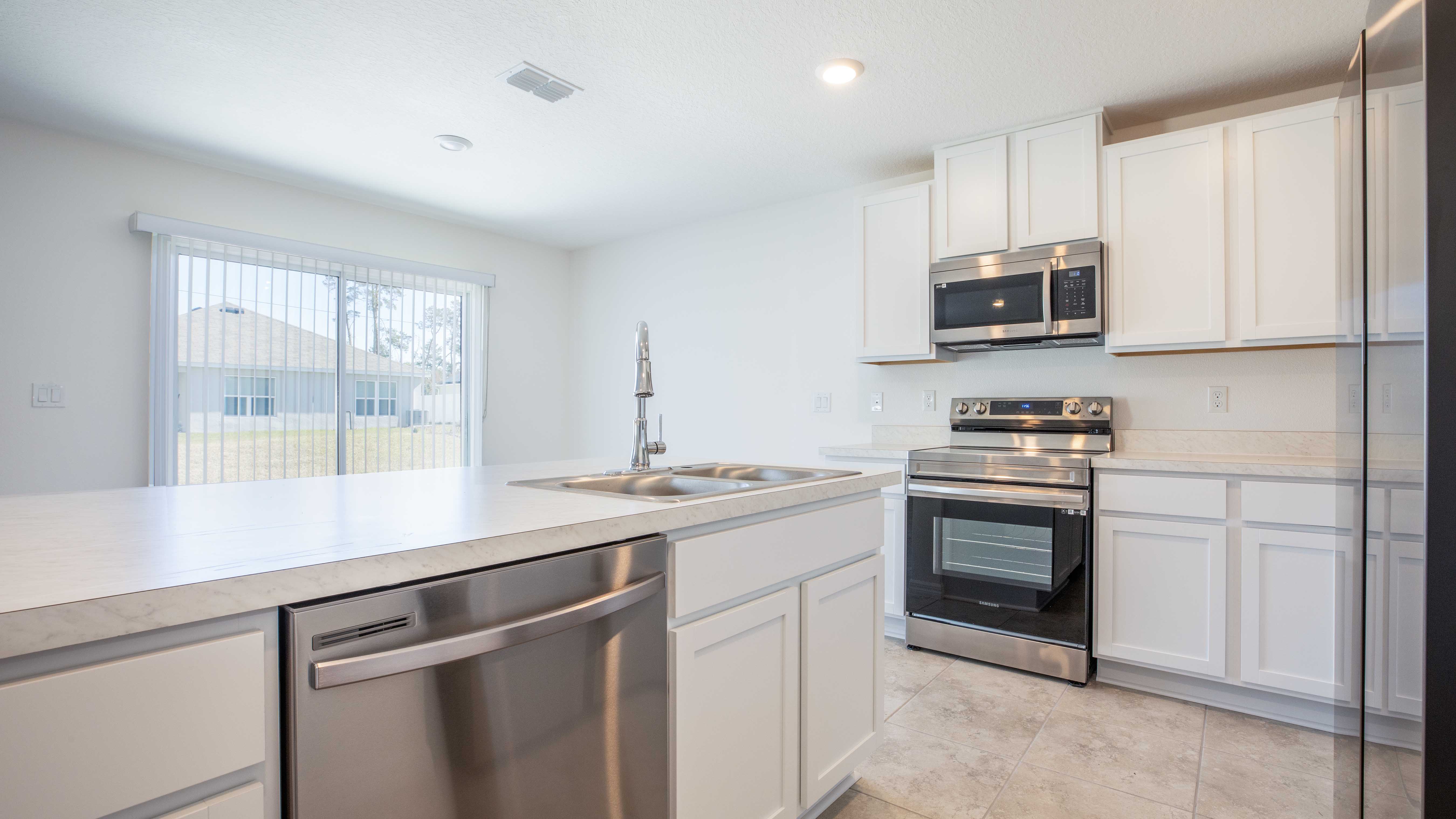 Modern kitchen with white cabinets, stainless steel appliances, and a large island with dual sink, illuminated by natural light from sliding glass doors.