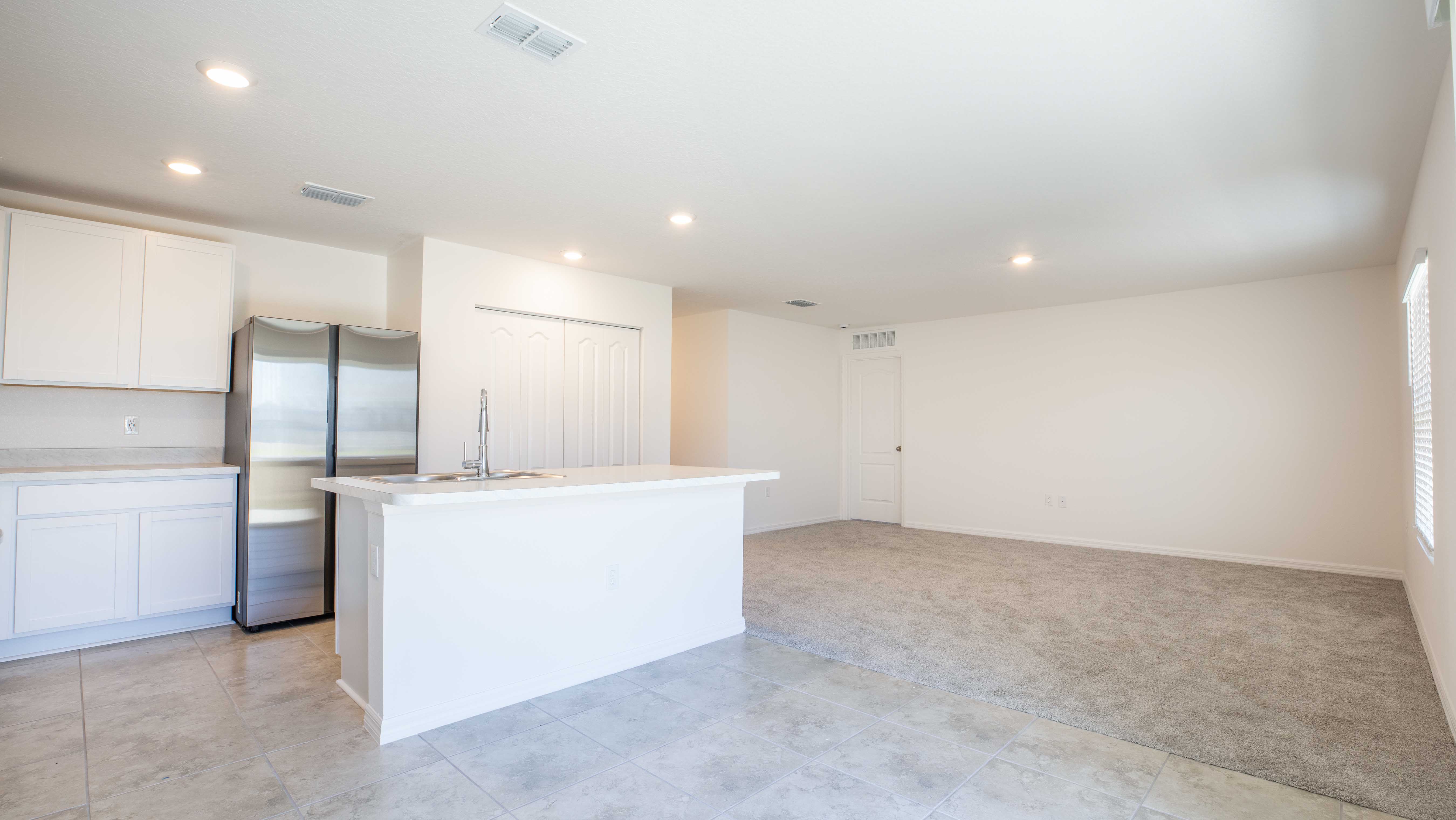 Modern open-concept kitchen and living area with white cabinets, stainless steel appliances, tiled flooring, and carpeted living space.