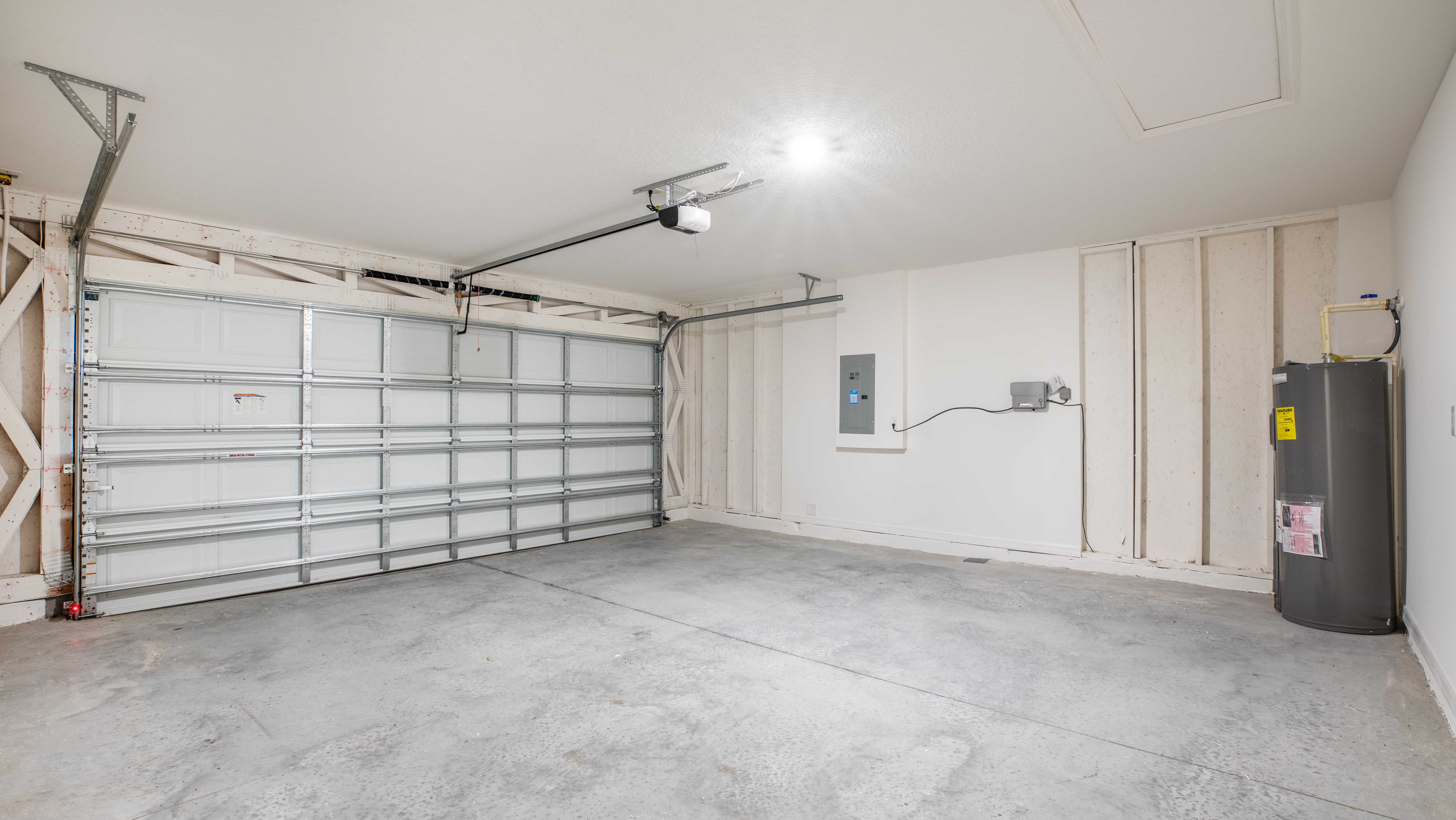 Empty two-car garage with concrete floors, white walls, and a closed roll-up door featuring a water heater and electrical panel on the side.