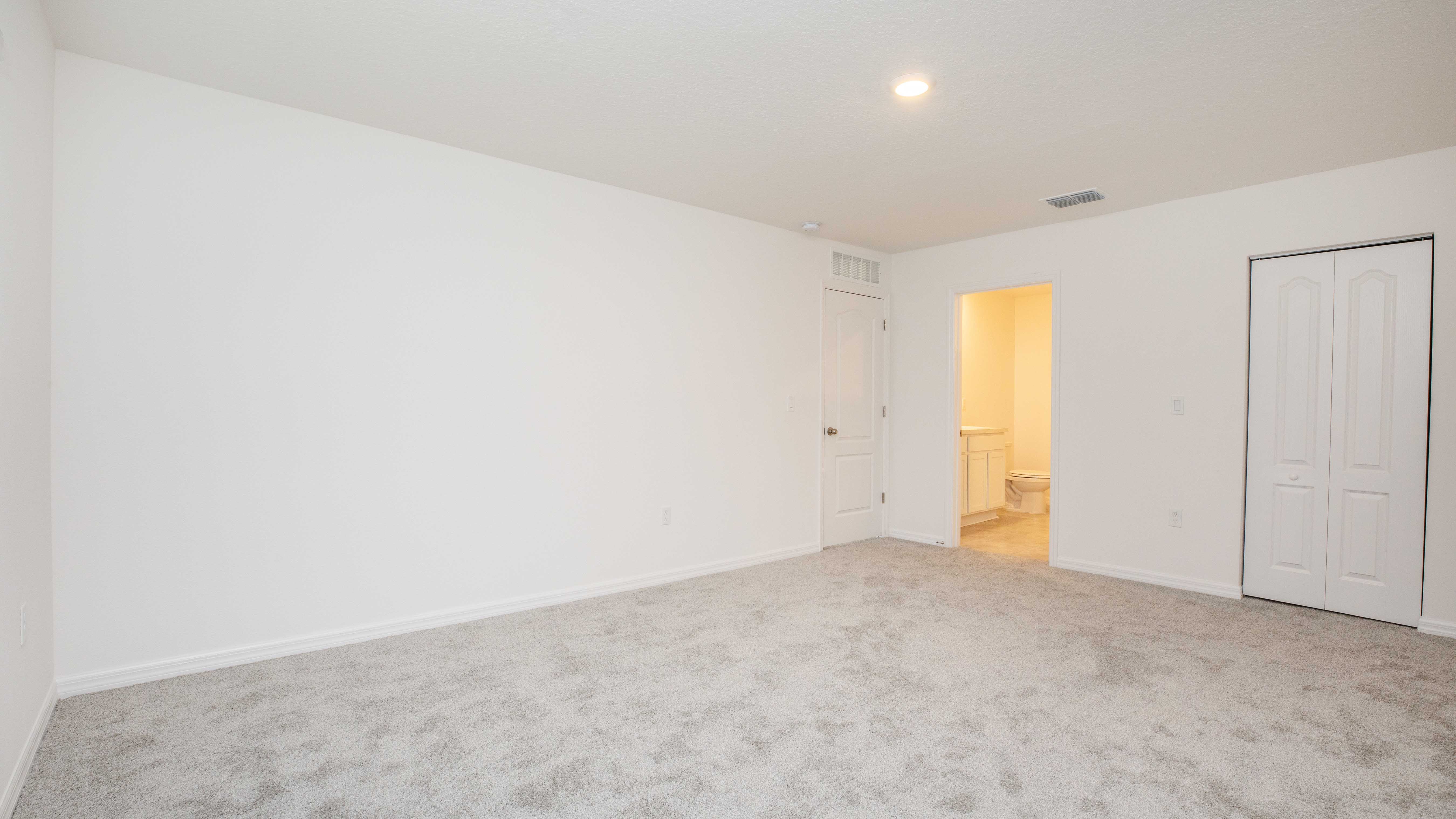 Empty carpeted room with white walls, a closet, and an open door leading to a lit bathroom.