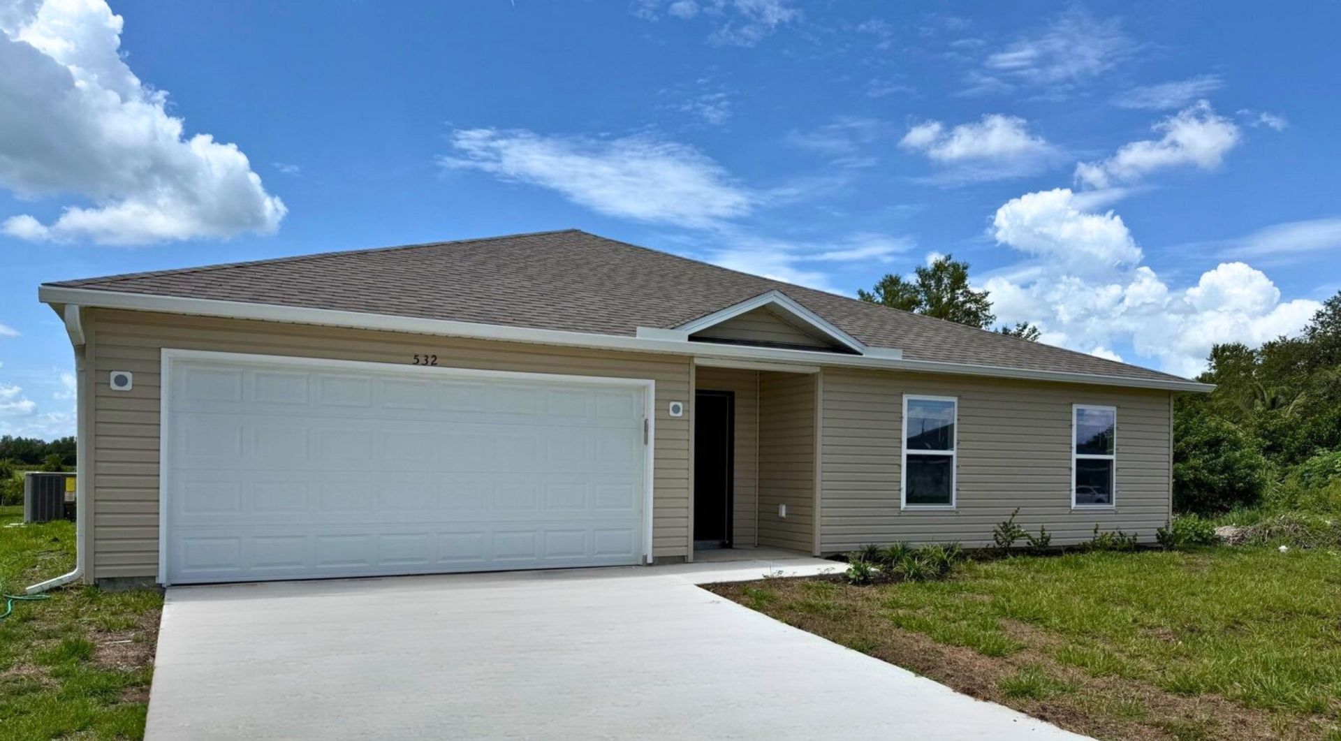 Single-story beige house with a two-car garage and well-maintained front lawn under a clear blue sky.