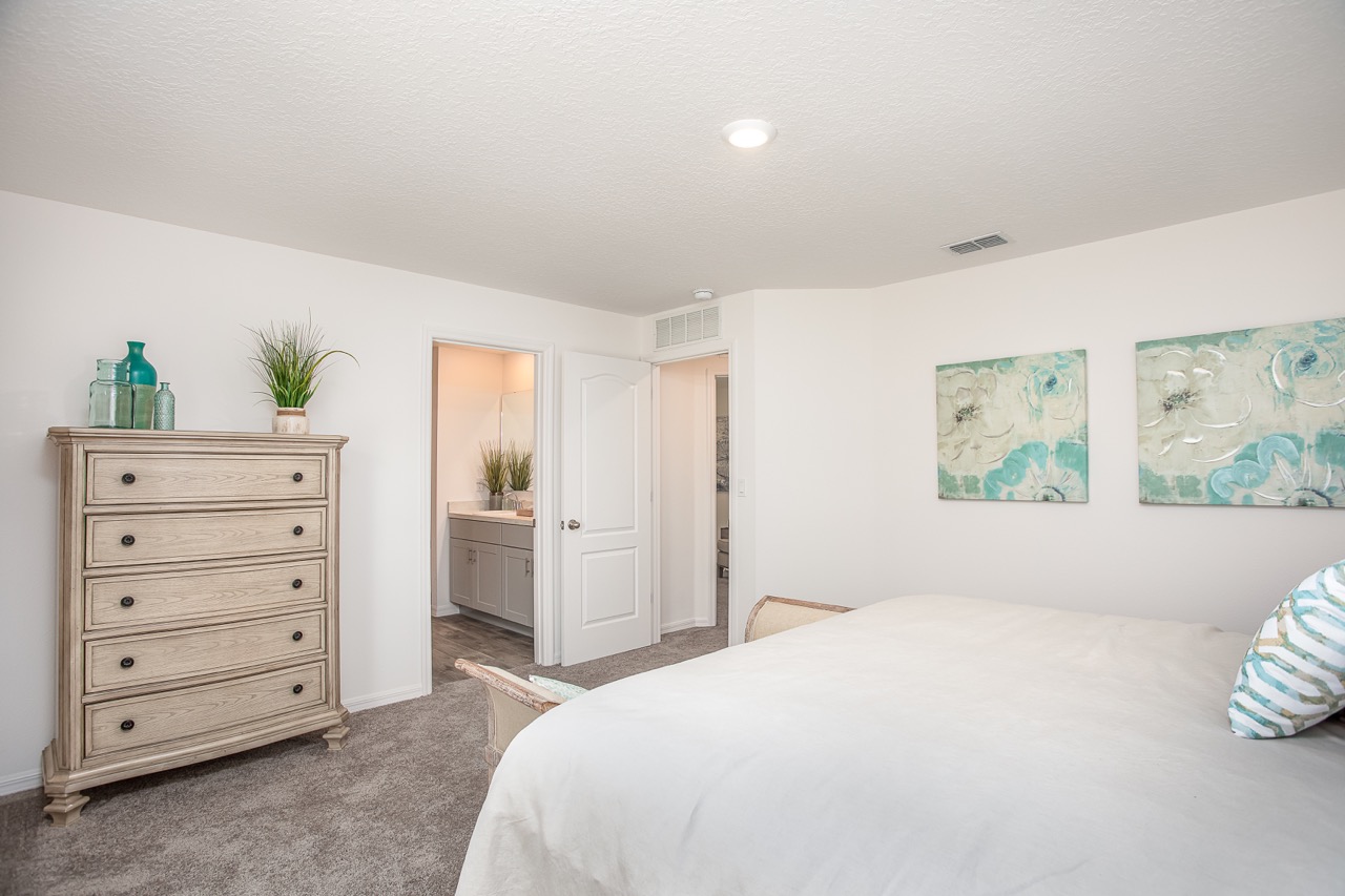 Modern bedroom with neutral decor, featuring a wooden dresser, wall art, and a view into a well-lit bathroom.
