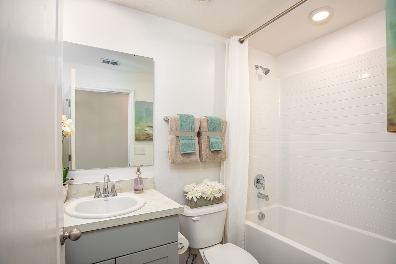 Stylish modern bathroom with white tiles, a vanity mirror, sink, and shower, accented by green and beige towels and decorative flowers.