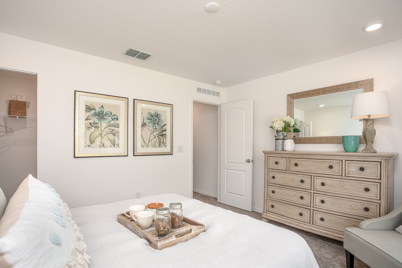 "Inviting modern bedroom with white decor, featuring framed botanical prints, a wooden dresser with a mirror, and a cozy coffee tray on the bed."