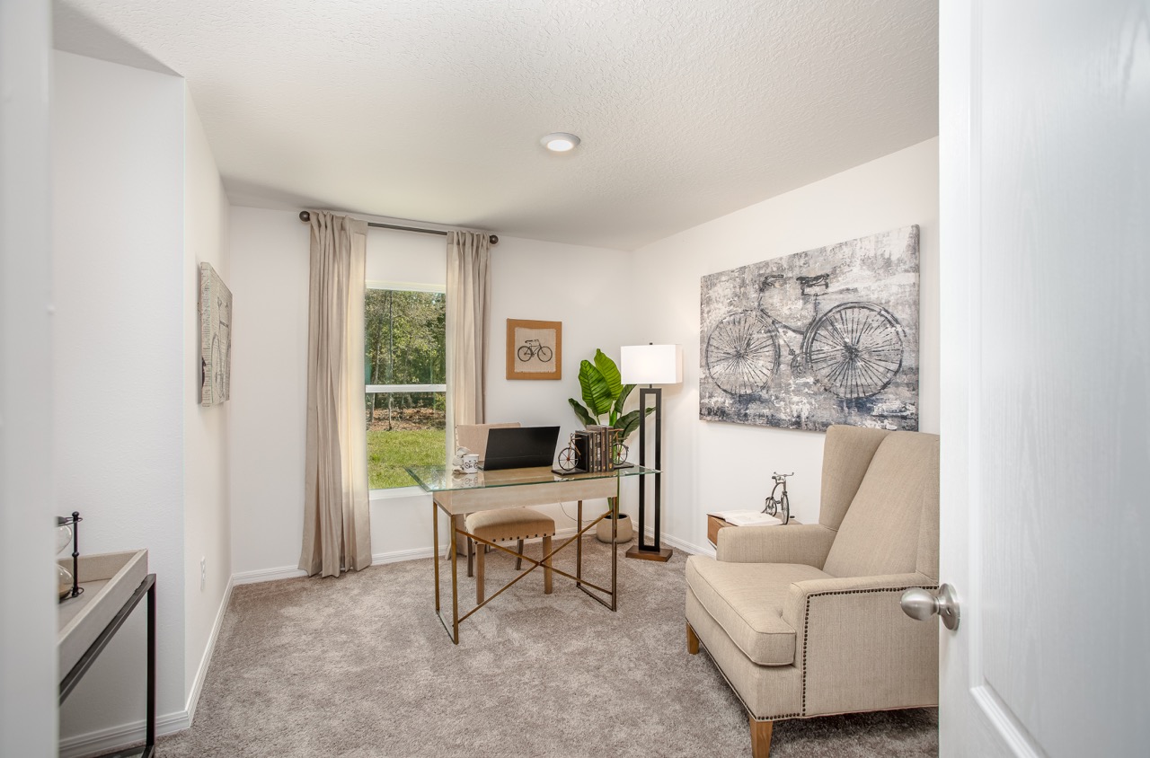 A modern home office with a glass desk, beige armchair, bicycle artwork, and a large window offering natural light.