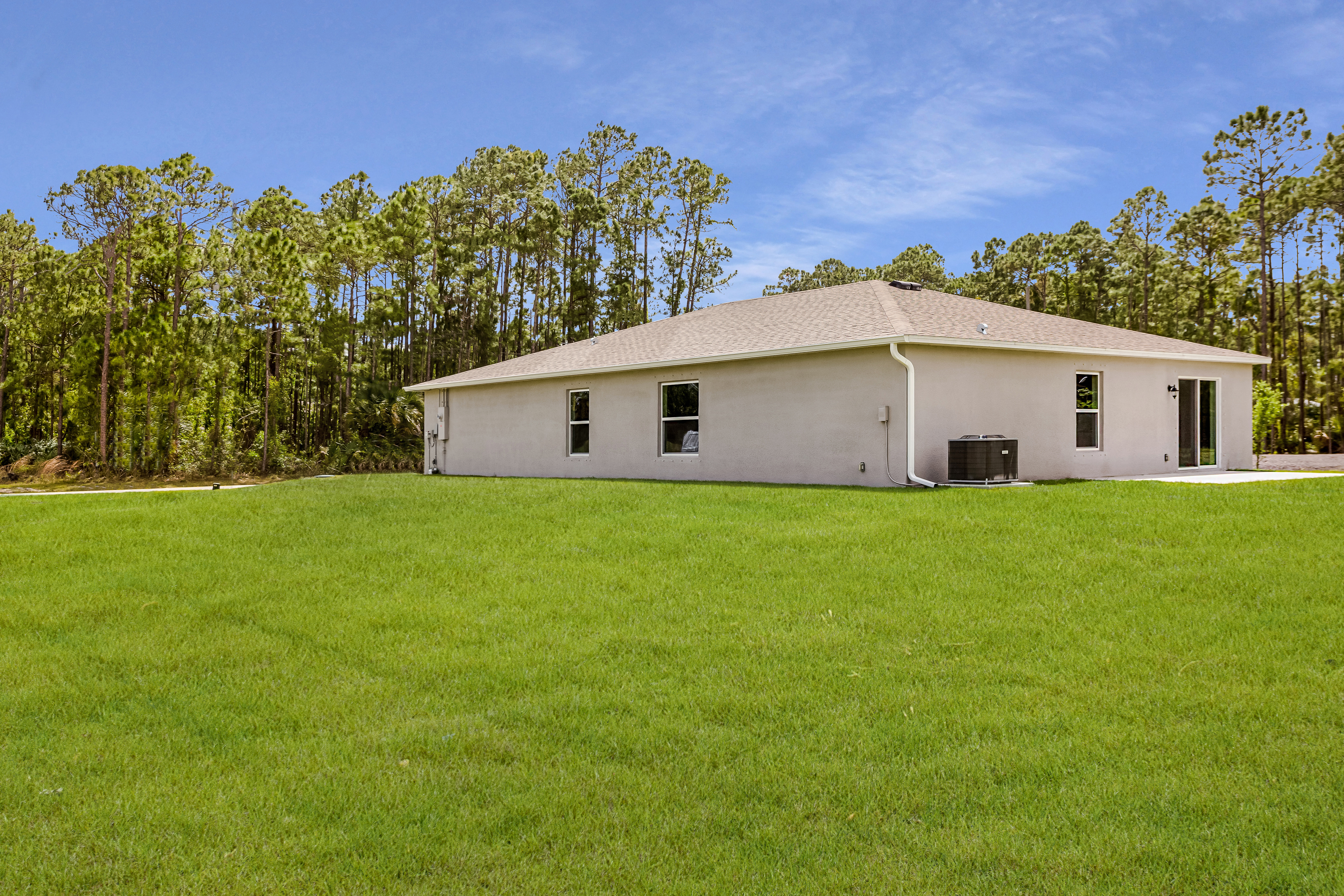 Single-story beige house with a sloped roof, surrounded by lush green grass and a backdrop of tall trees, under a clear blue sky.