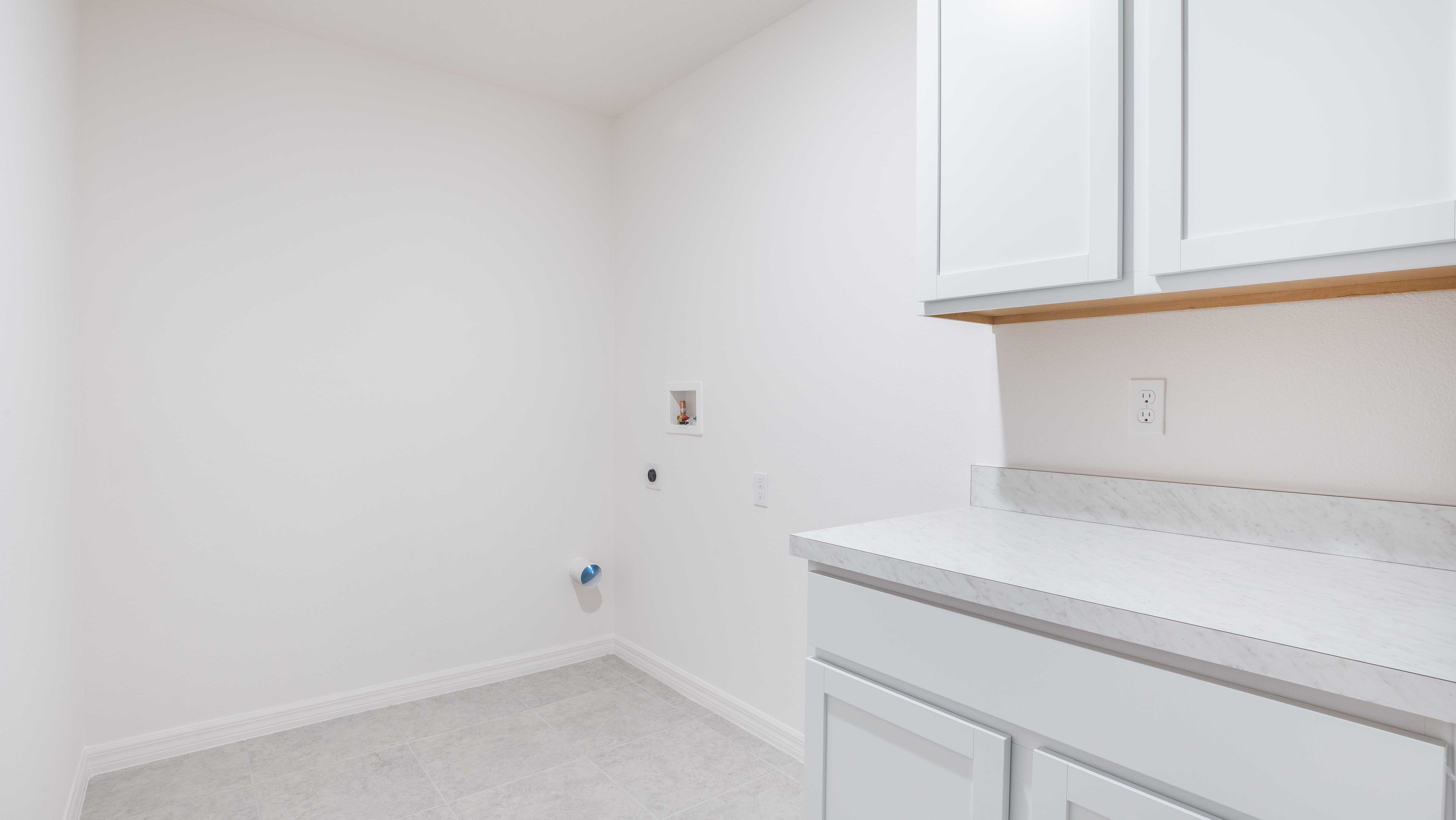 Minimalist laundry room with white cabinets, countertop, and tile floor, featuring plumbing and electrical connections.