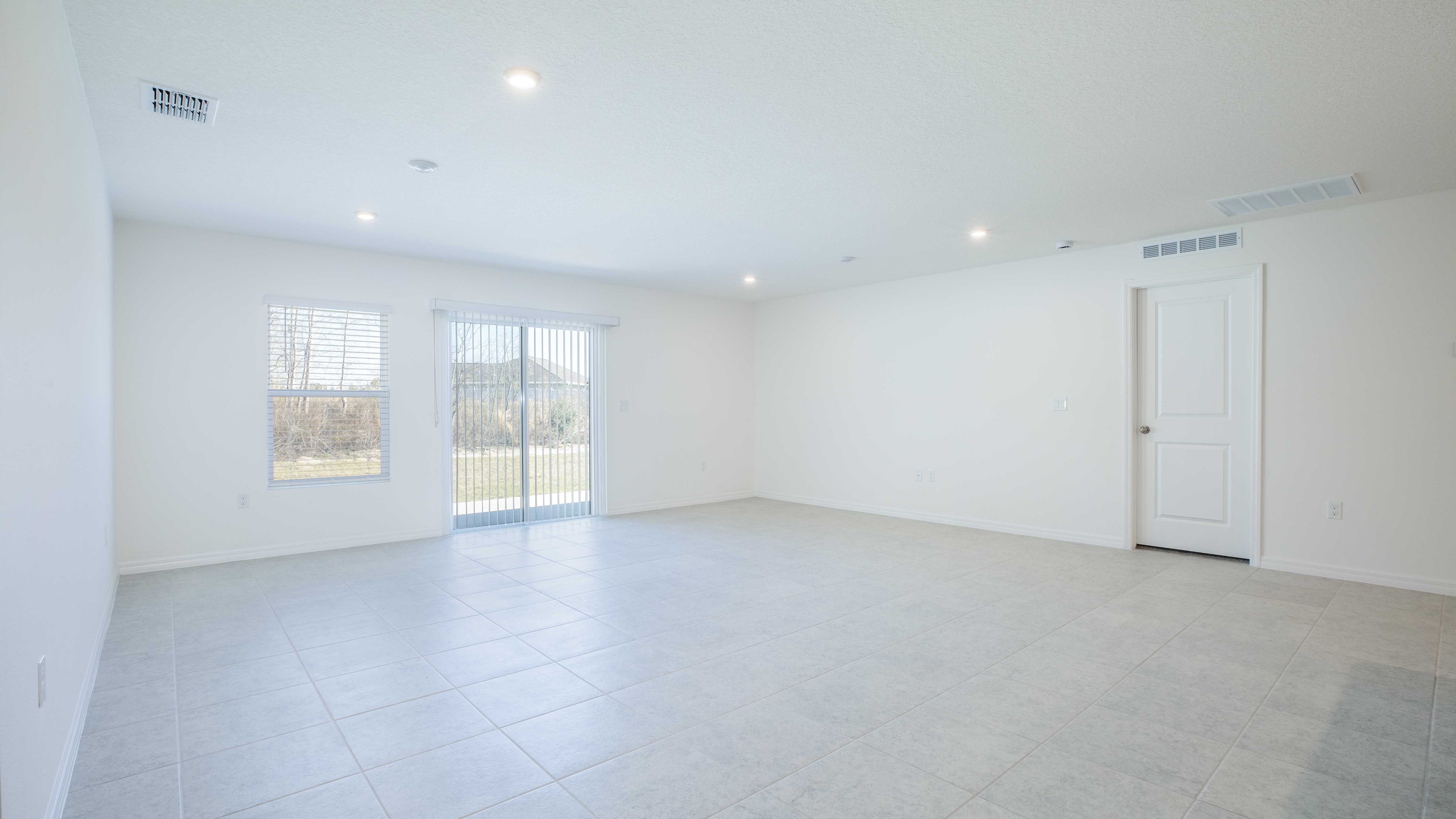 Spacious, empty living room with large tile flooring, white walls, a door, and sliding glass patio doors.