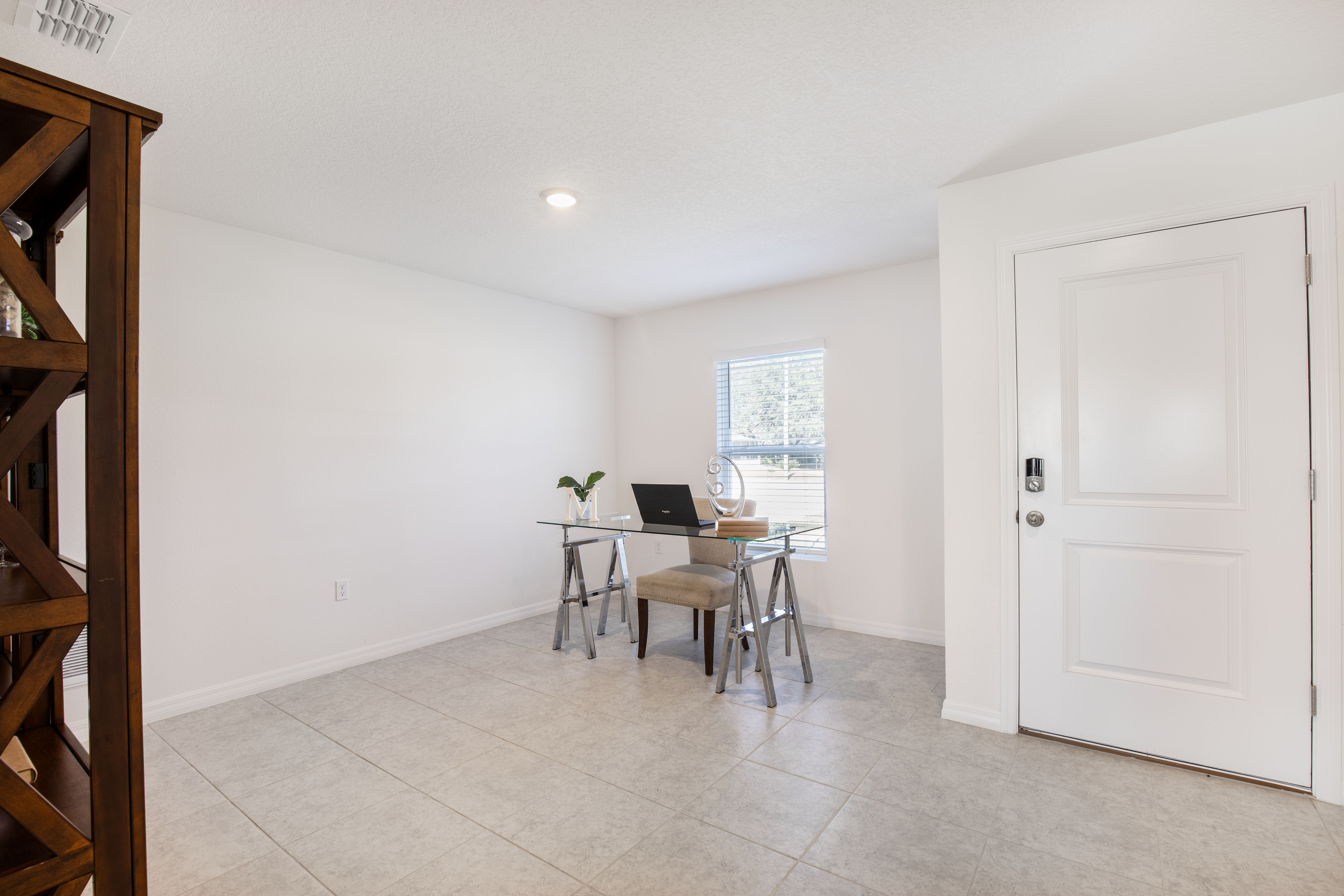 Minimalist home office with a glass desk, laptop, and beige chair near a window in a tiled room.