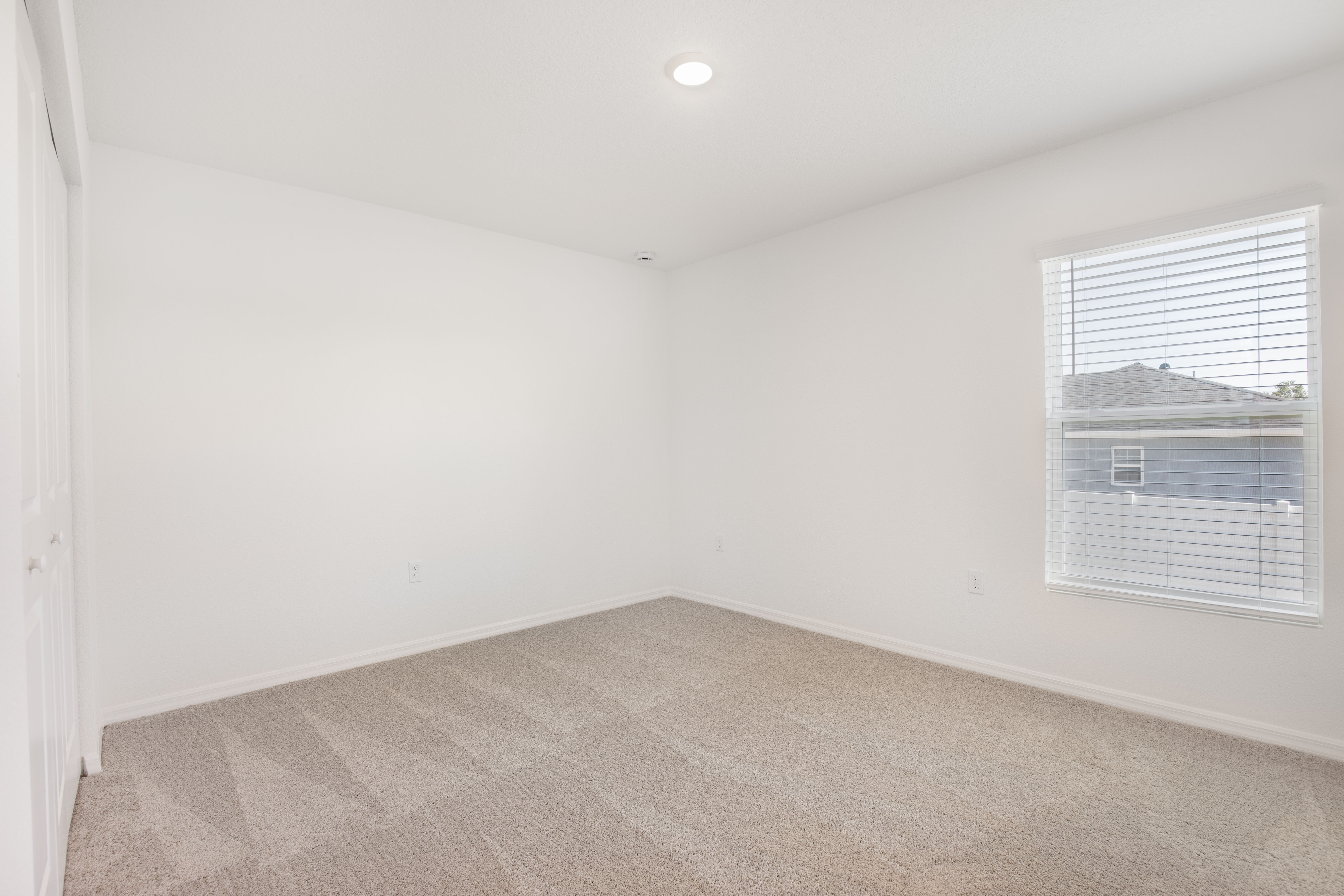 Empty room with beige carpet and white walls, featuring a single window with blinds and a ceiling light.