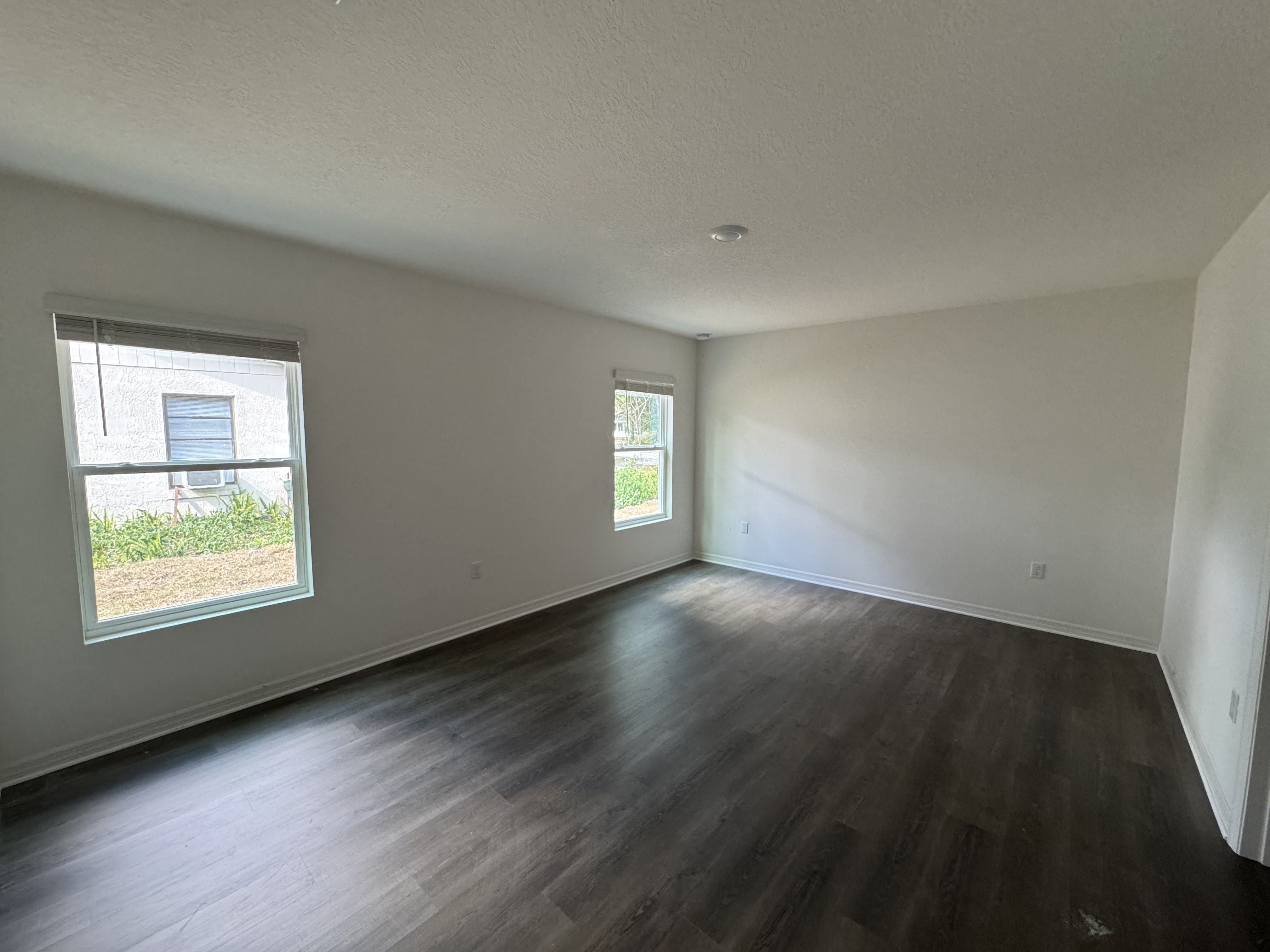 A spacious, unfurnished room with dark wooden floors and two large windows allowing natural light to brighten the white-painted walls.