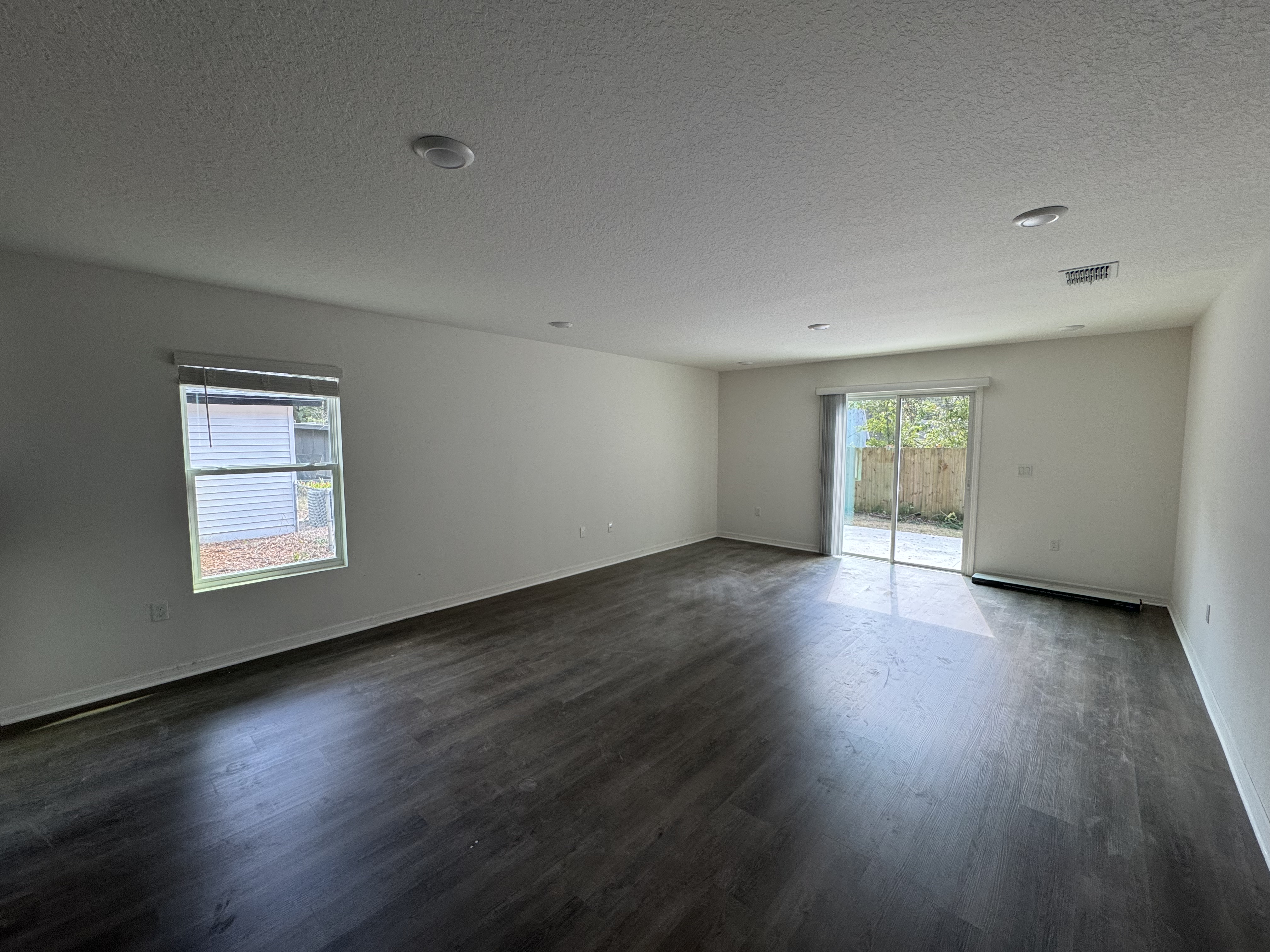 Spacious empty room with dark wood flooring, large window, and sliding glass door leading to a fenced backyard.