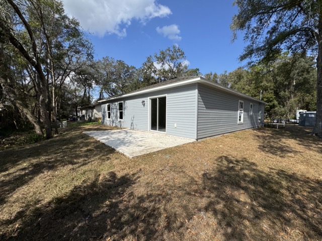 Single-story gray house with a sloped roof and patio, surrounded by trees and grassy yard.