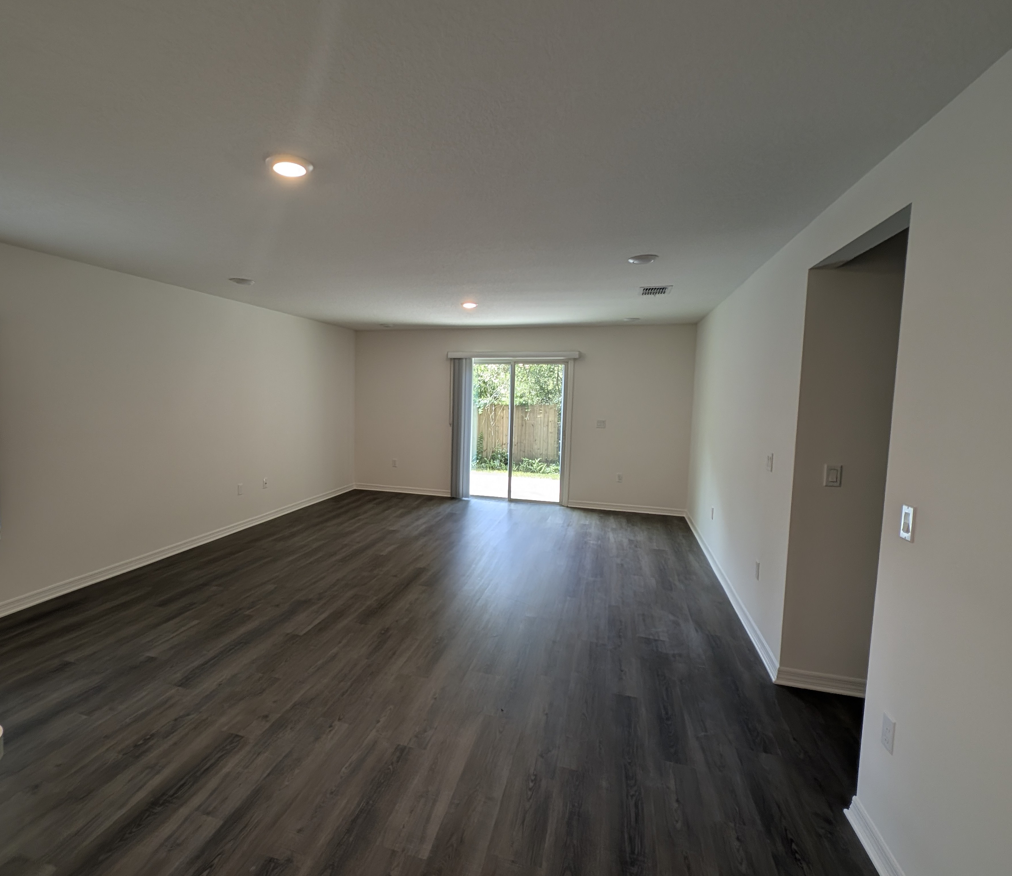 Spacious empty living room with dark wood flooring, recessed lighting, and a sliding glass door leading to a backyard.