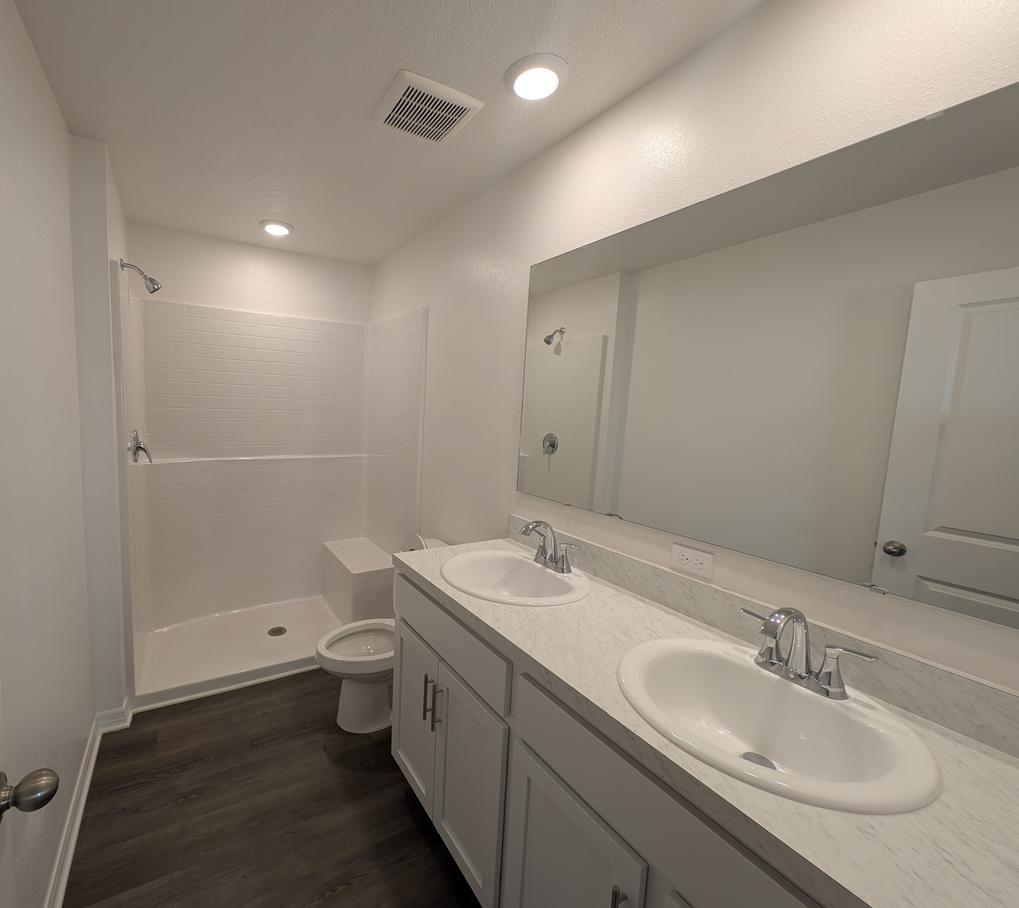 Modern bathroom with dual sinks, a large mirror, and a walk-in shower on wood-style flooring.