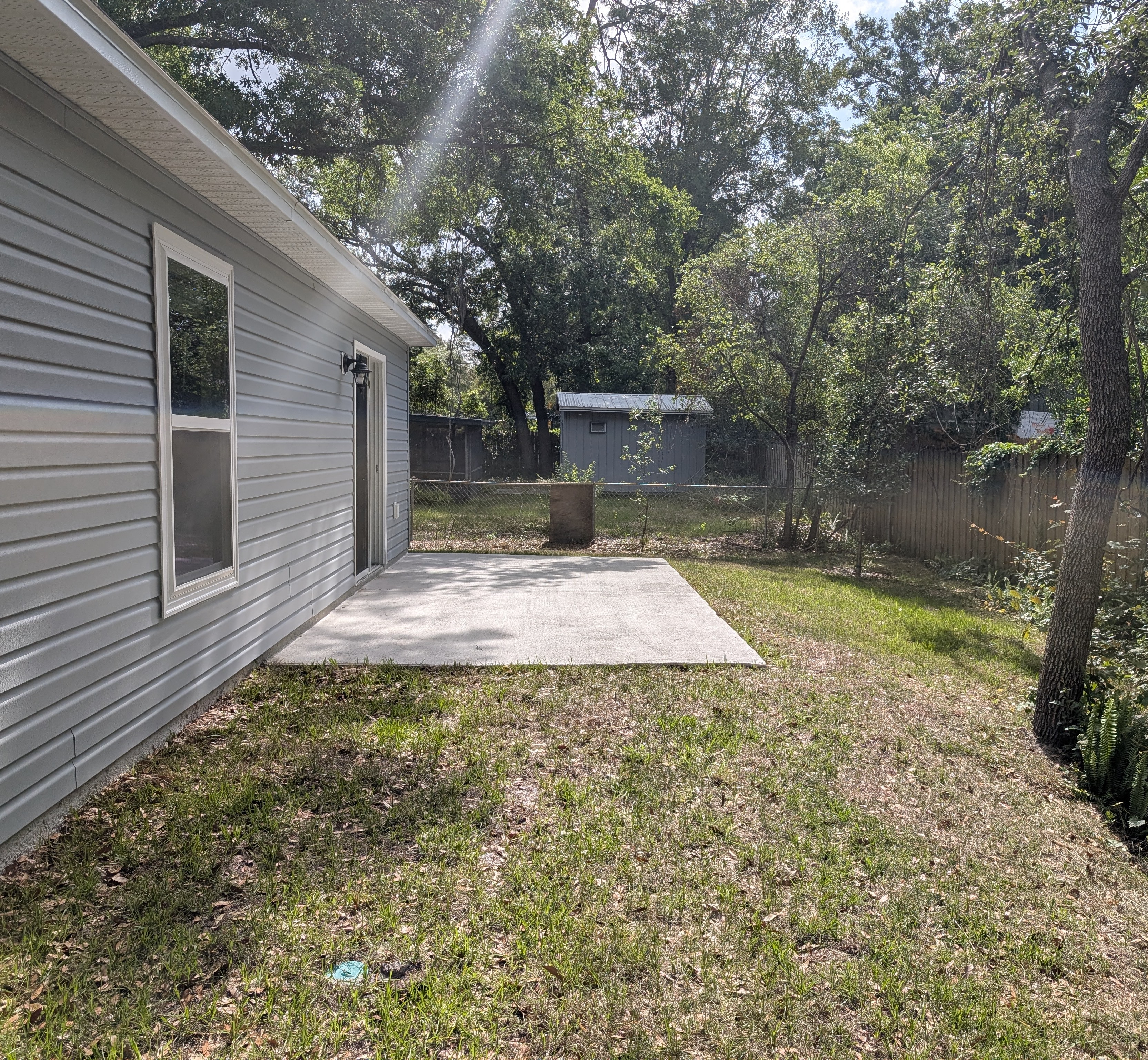 Backyard view of a house with a concrete patio, surrounded by greenery and a storage shed in the distance.