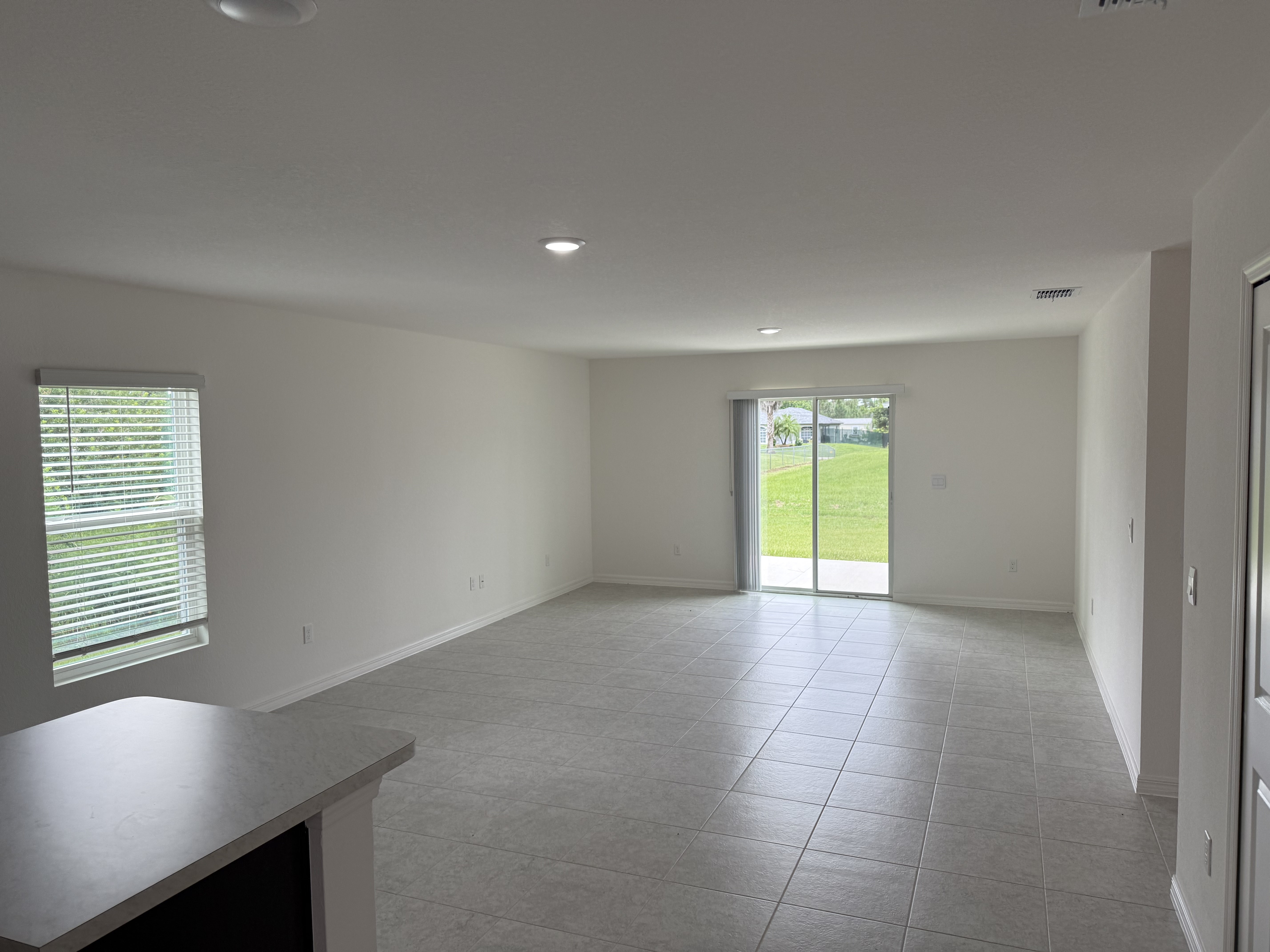Modern empty living room with white walls, large window, sliding glass door, and tiled flooring overlooking a grassy yard.