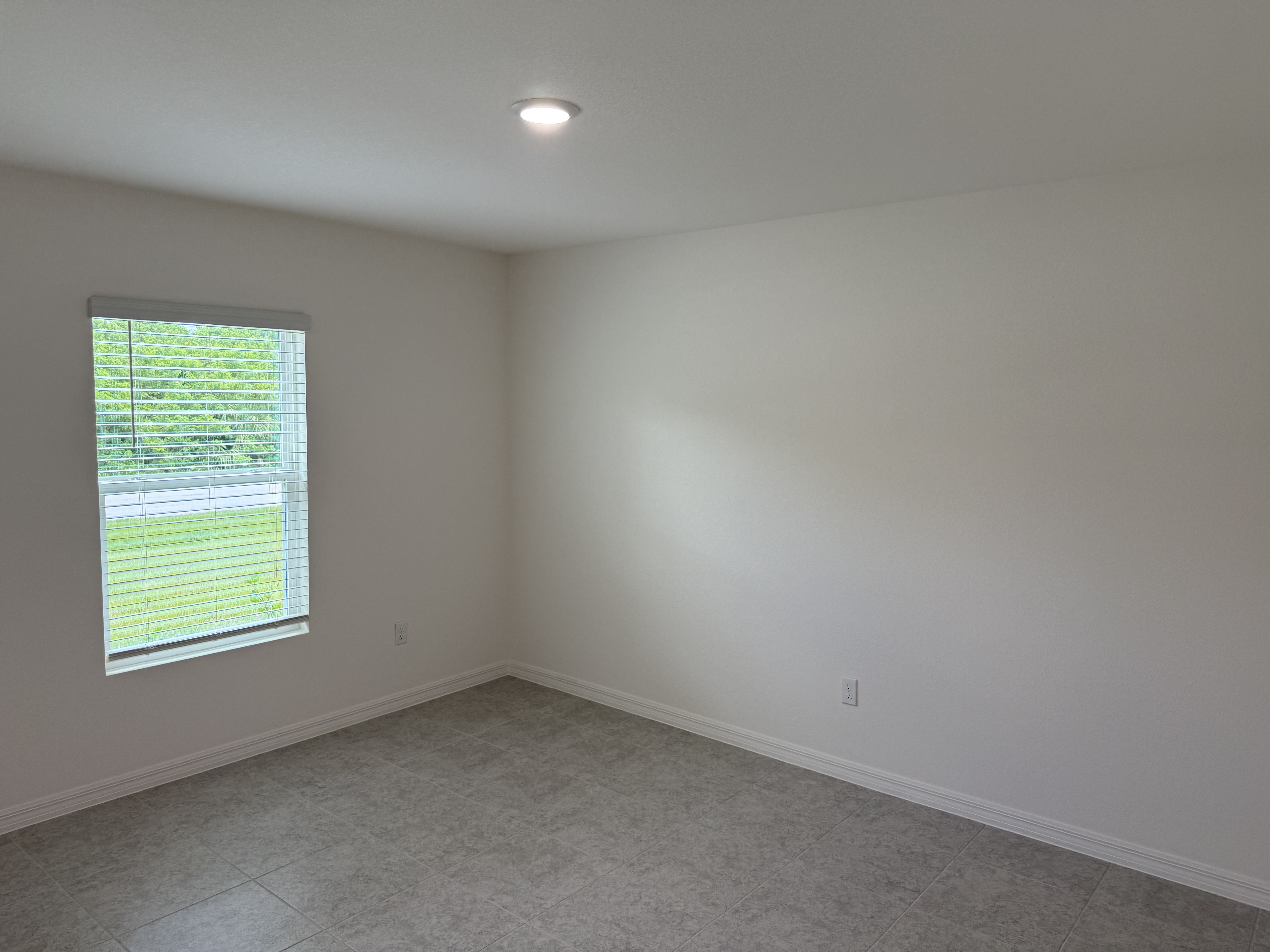 Empty room with white walls, a single window with blinds, and tiled flooring, illuminated by ceiling light.