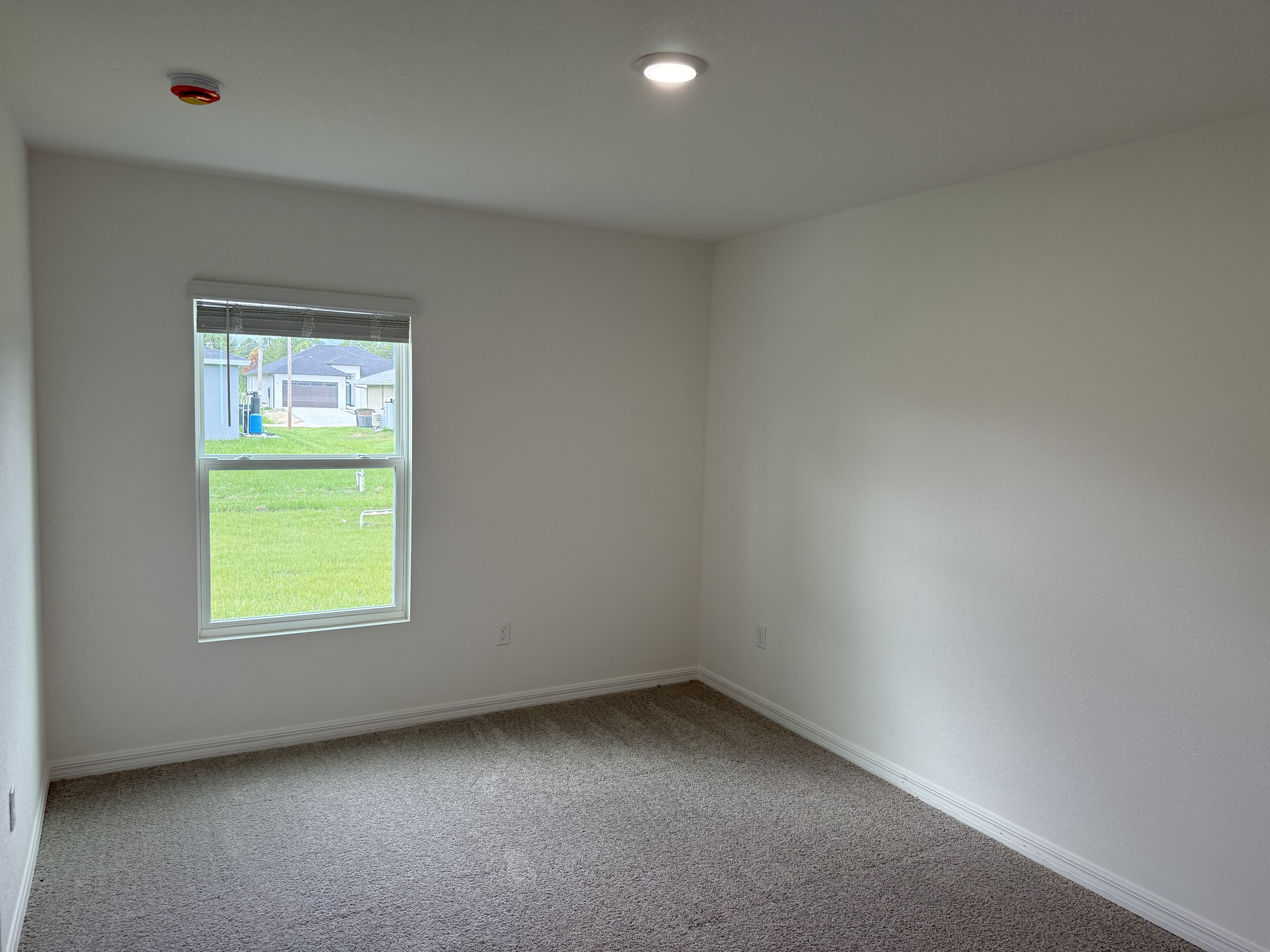 Empty room with beige carpet, white walls, and a window showing a view of grass and neighboring houses.