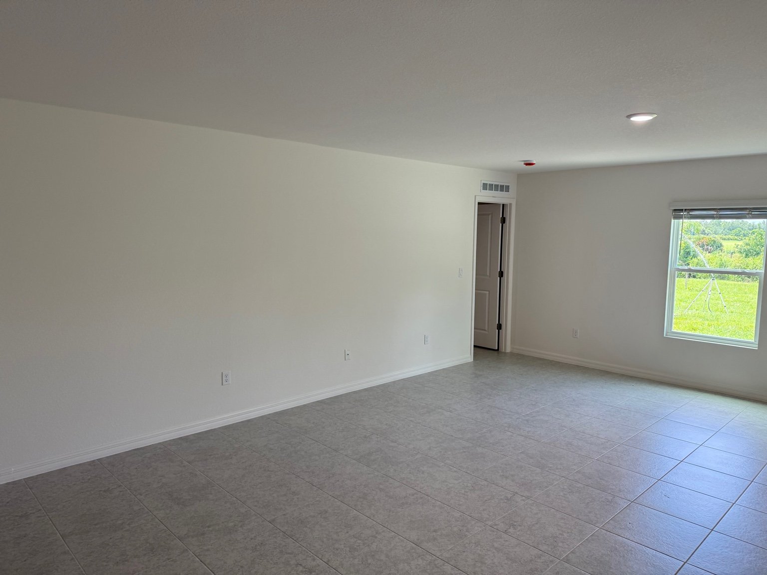 Spacious empty room with tile flooring, white walls, and a large window offering a view of green grass outside.