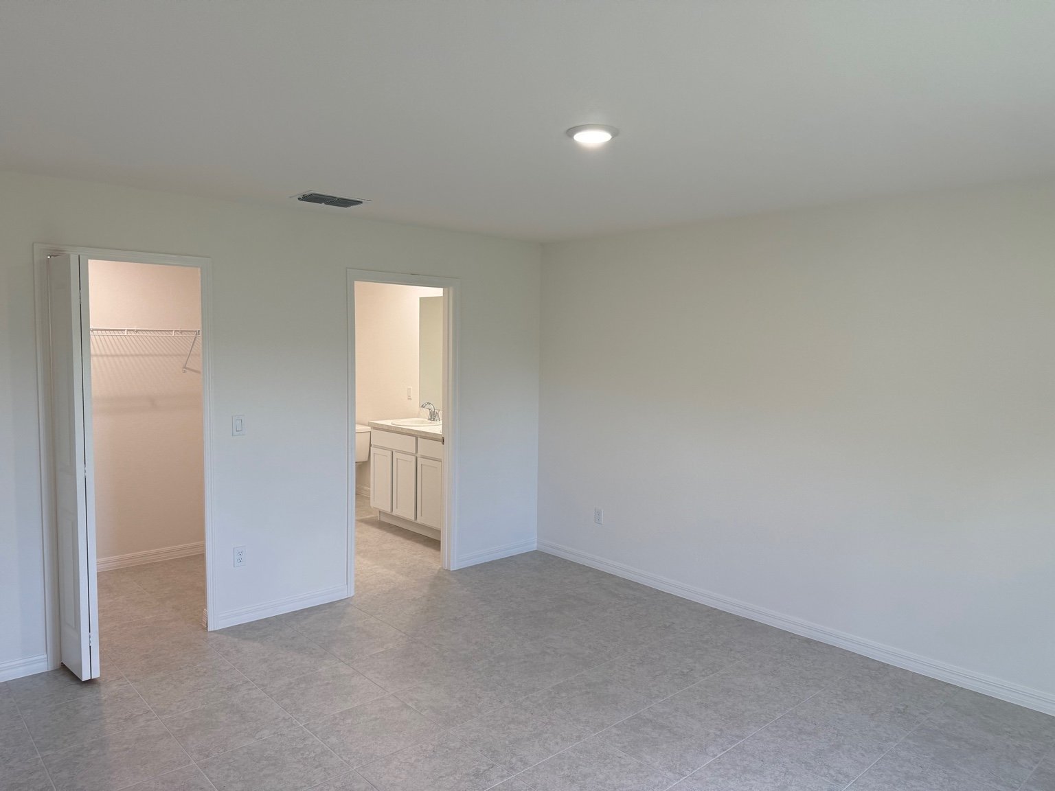 An empty, newly renovated room with tiled flooring, featuring an open door to a closet and another to a bathroom with a white vanity.
