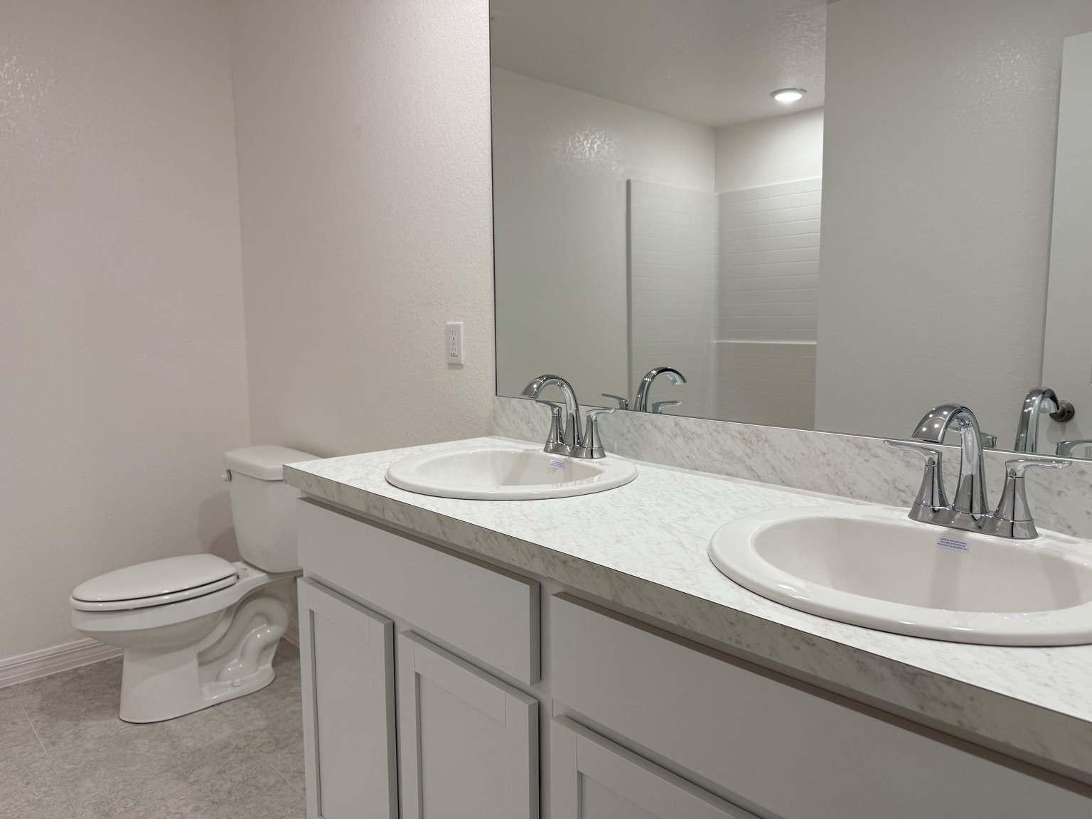 Modern bathroom with dual sinks, marble countertop, white cabinetry, and a view of a toilet and shower area.