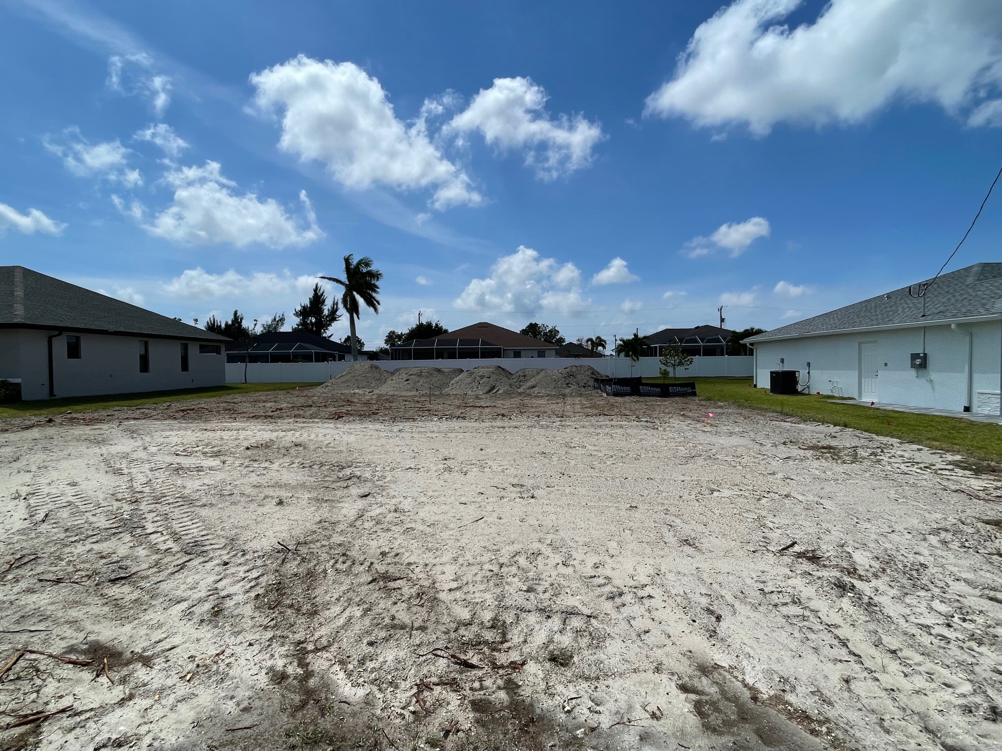Vacant residential plot with dirt mounds, surrounded by houses on a sunny day.