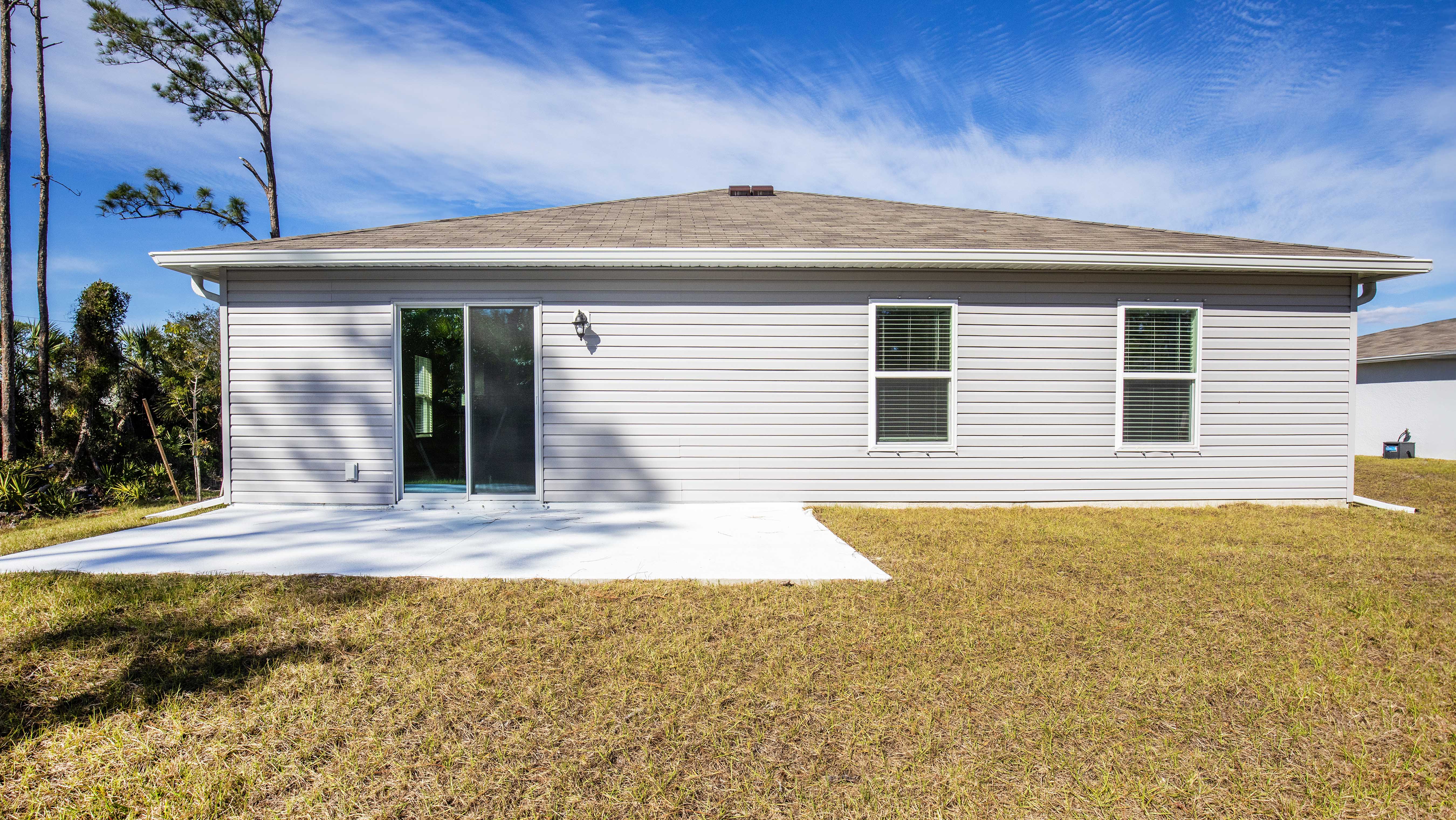 The image showcases the back view of a light gray house with a sliding glass door and two windows, set against a backdrop of trees and a blue sky.