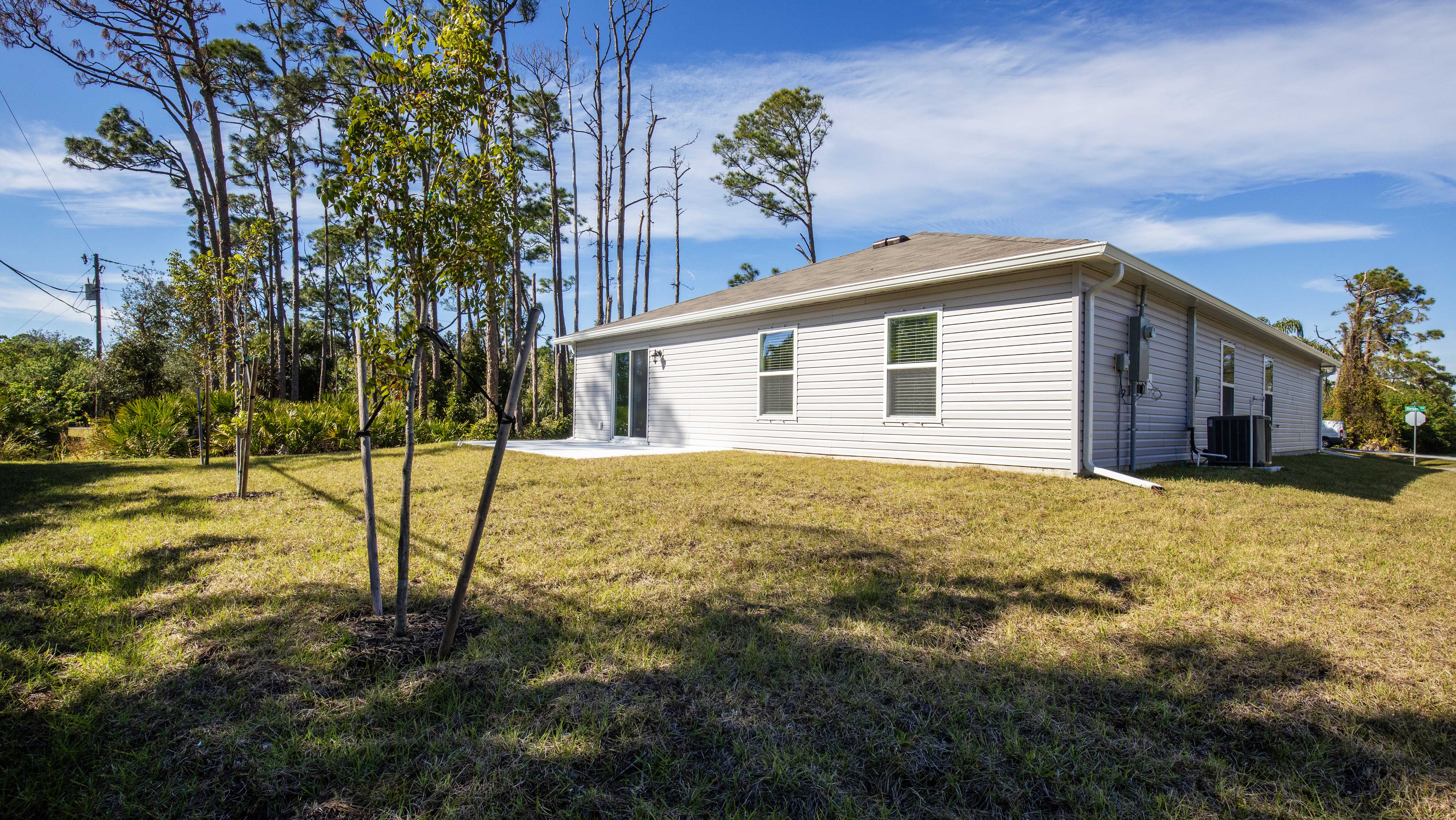 Single-story house with white siding in a grassy yard surrounded by tall trees and a clear blue sky.