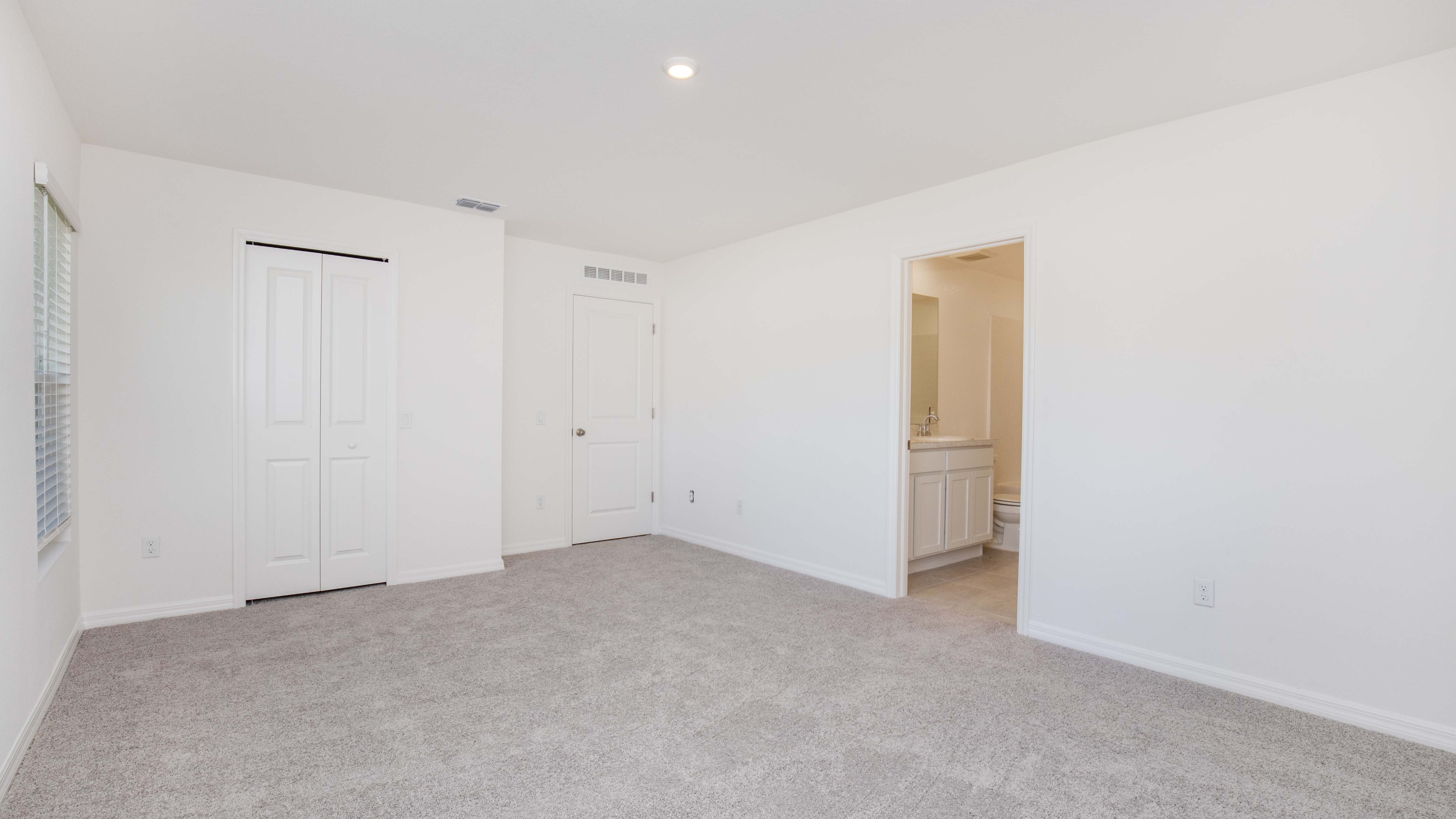 Bright, empty bedroom with light gray carpet, white walls, a closet, and a view into a bathroom.