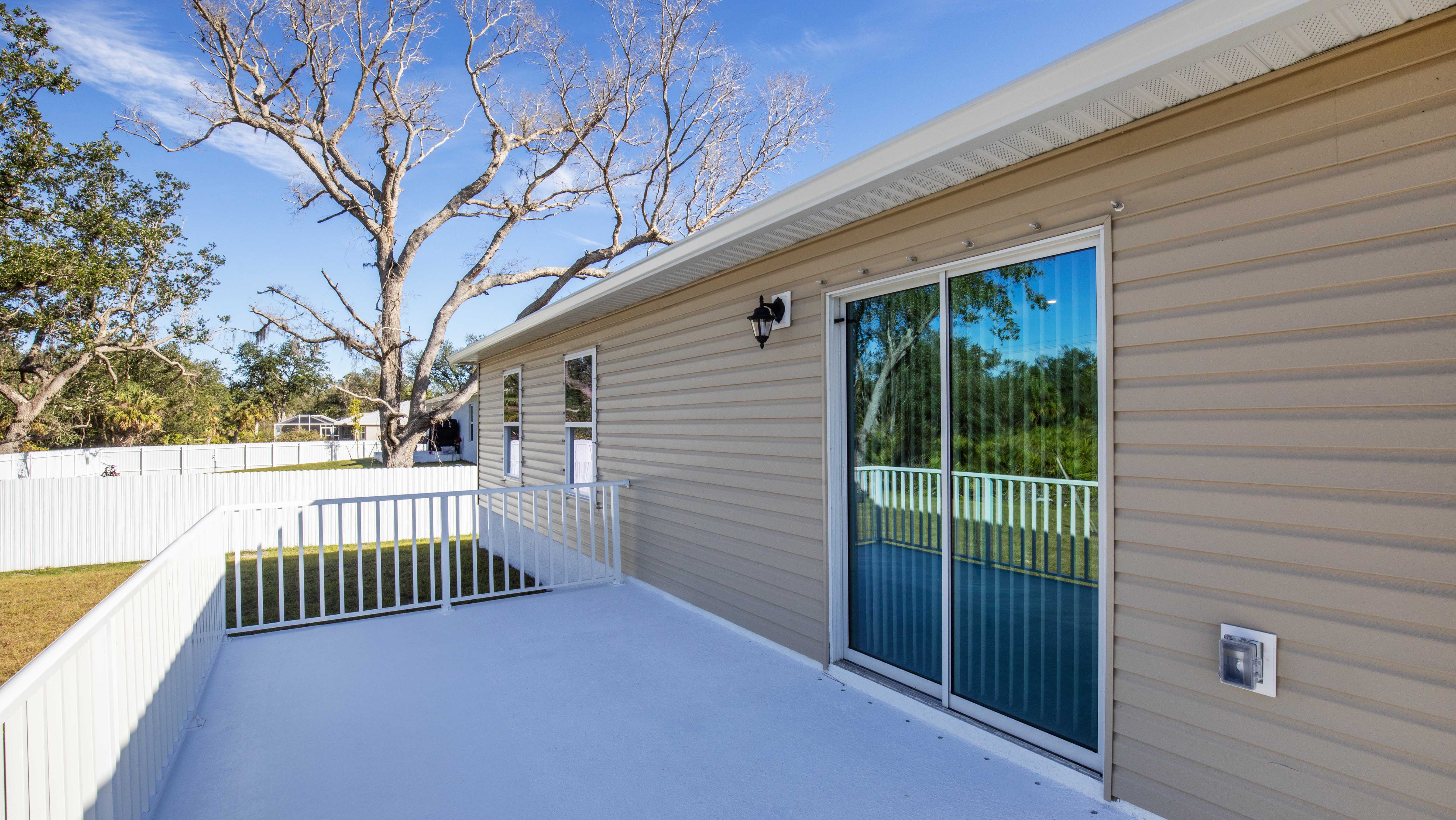 Elevated view of a beige house exterior with sliding glass doors opening to a white fenced patio, surrounded by trees and a blue sky.
