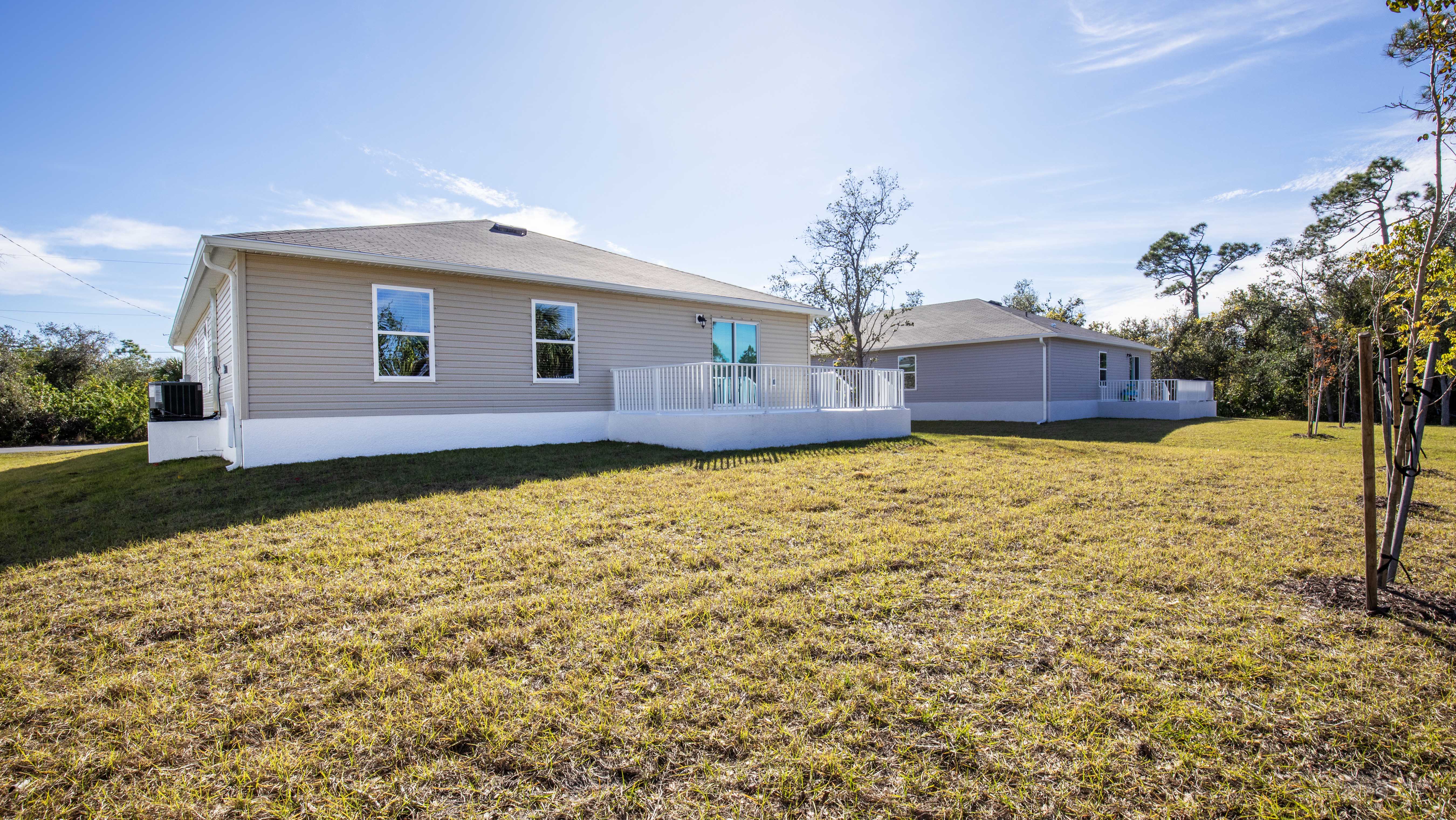 Two beige single-story homes with white railings and large grassy backyards under a clear blue sky.