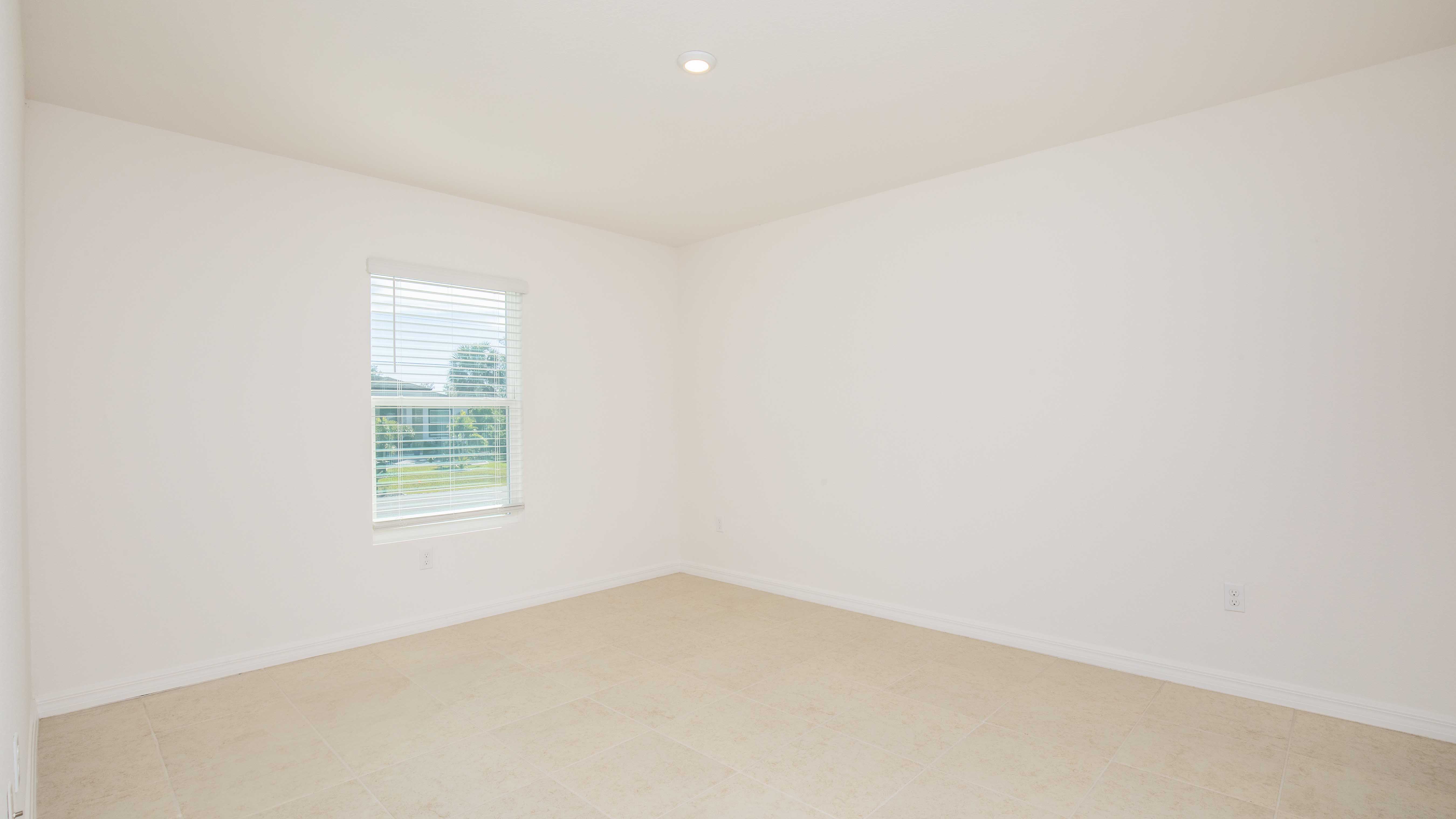 Empty, sunlit room with beige tiled floor, white walls, and a window showcasing greenery outside.