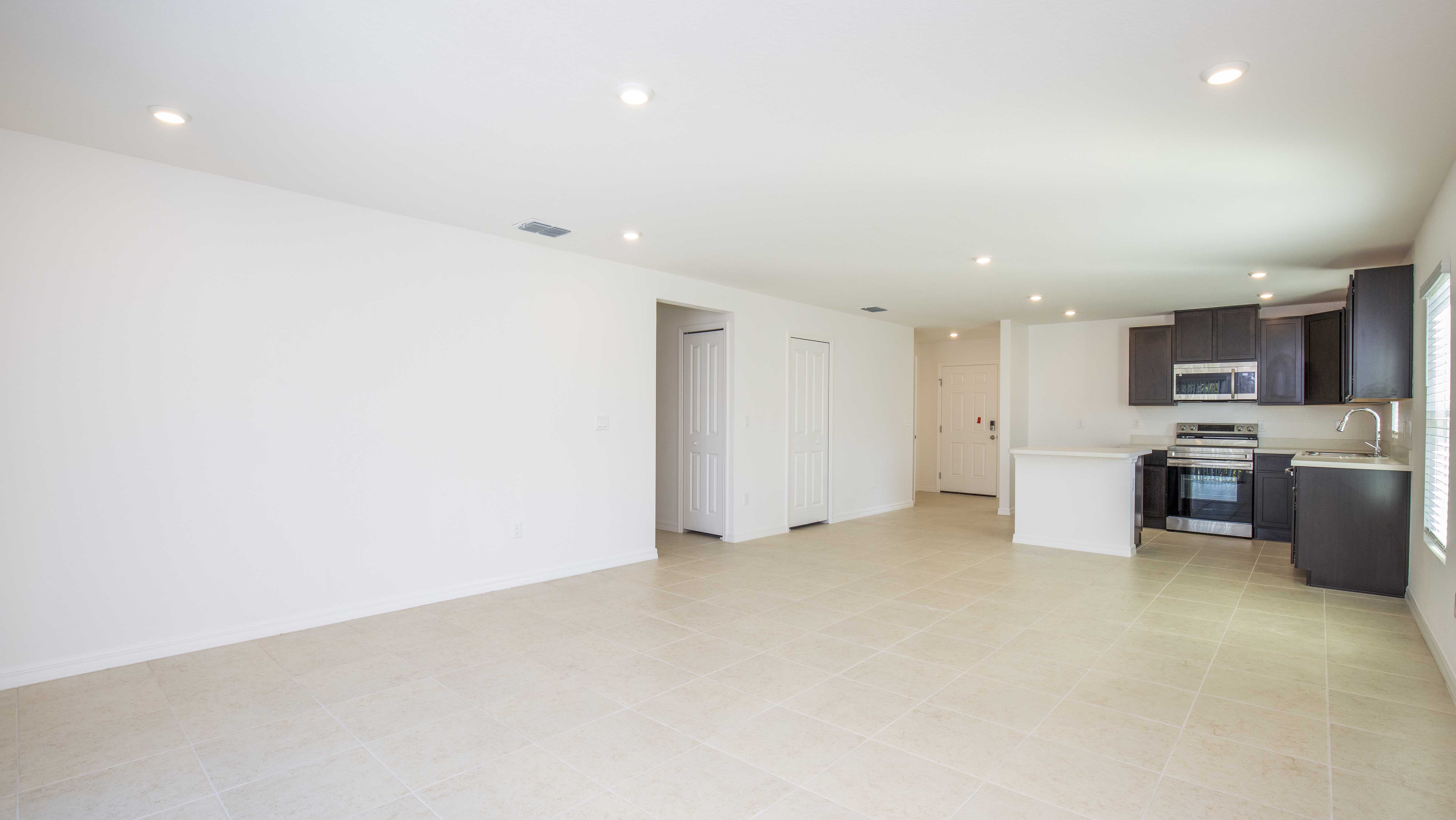 Bright, empty open-plan living room with modern kitchen, light tiled floors, and recessed lighting.