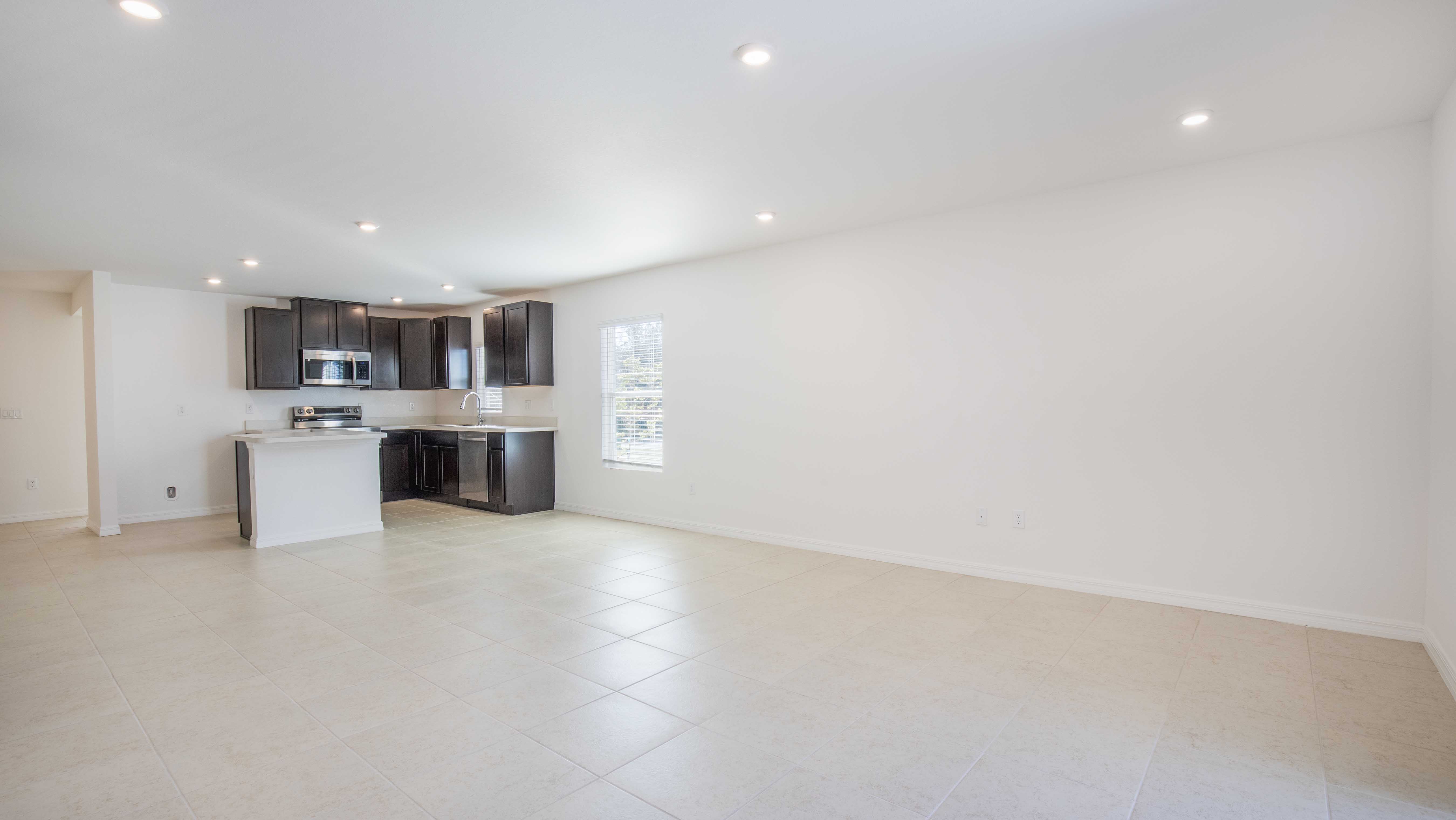 Modern open-plan kitchen with dark cabinets, stainless steel appliances, and white tiled flooring in a bright, spacious room.