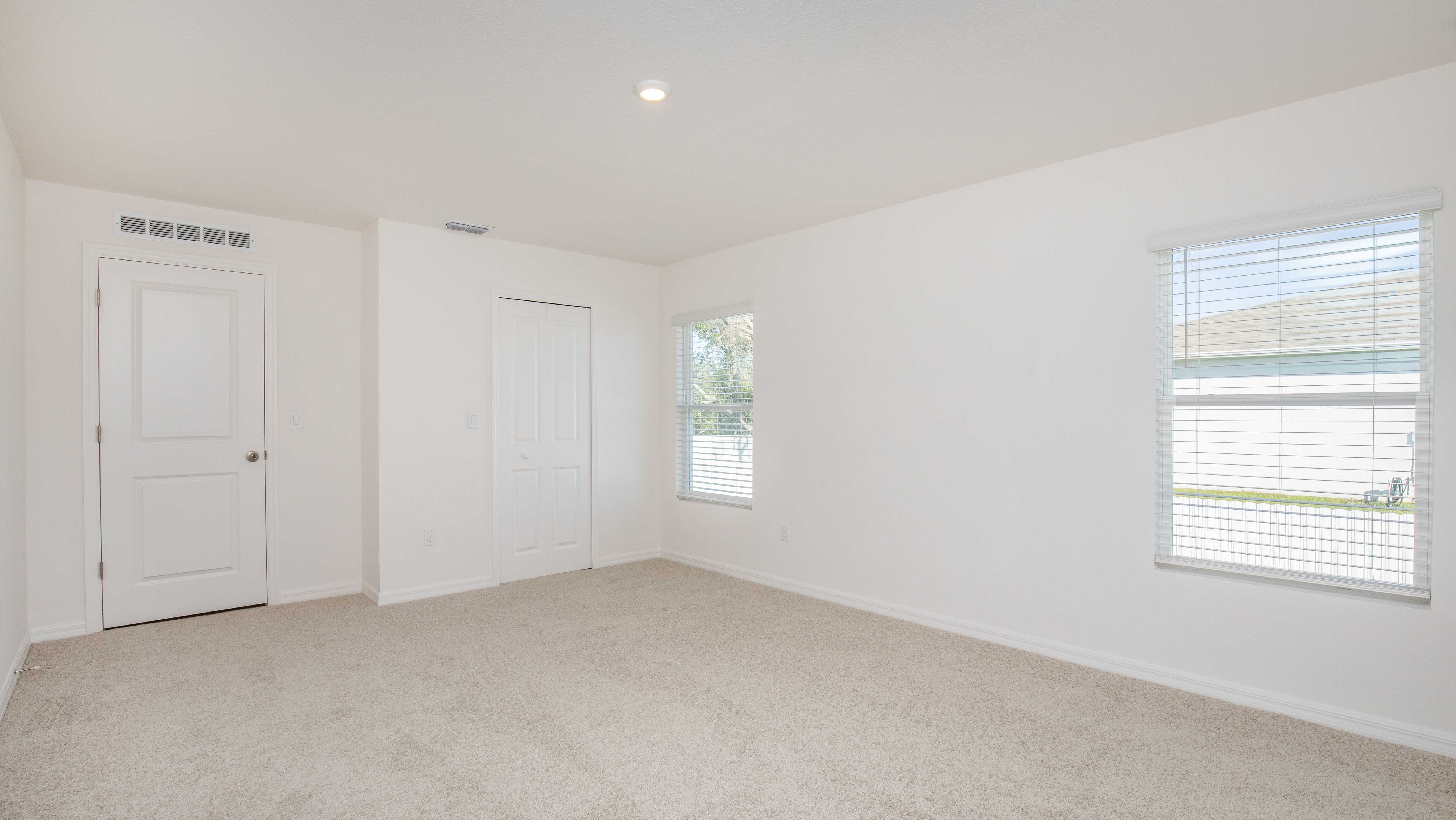 A bright, empty room with white walls, beige carpet, and large windows allowing natural light.