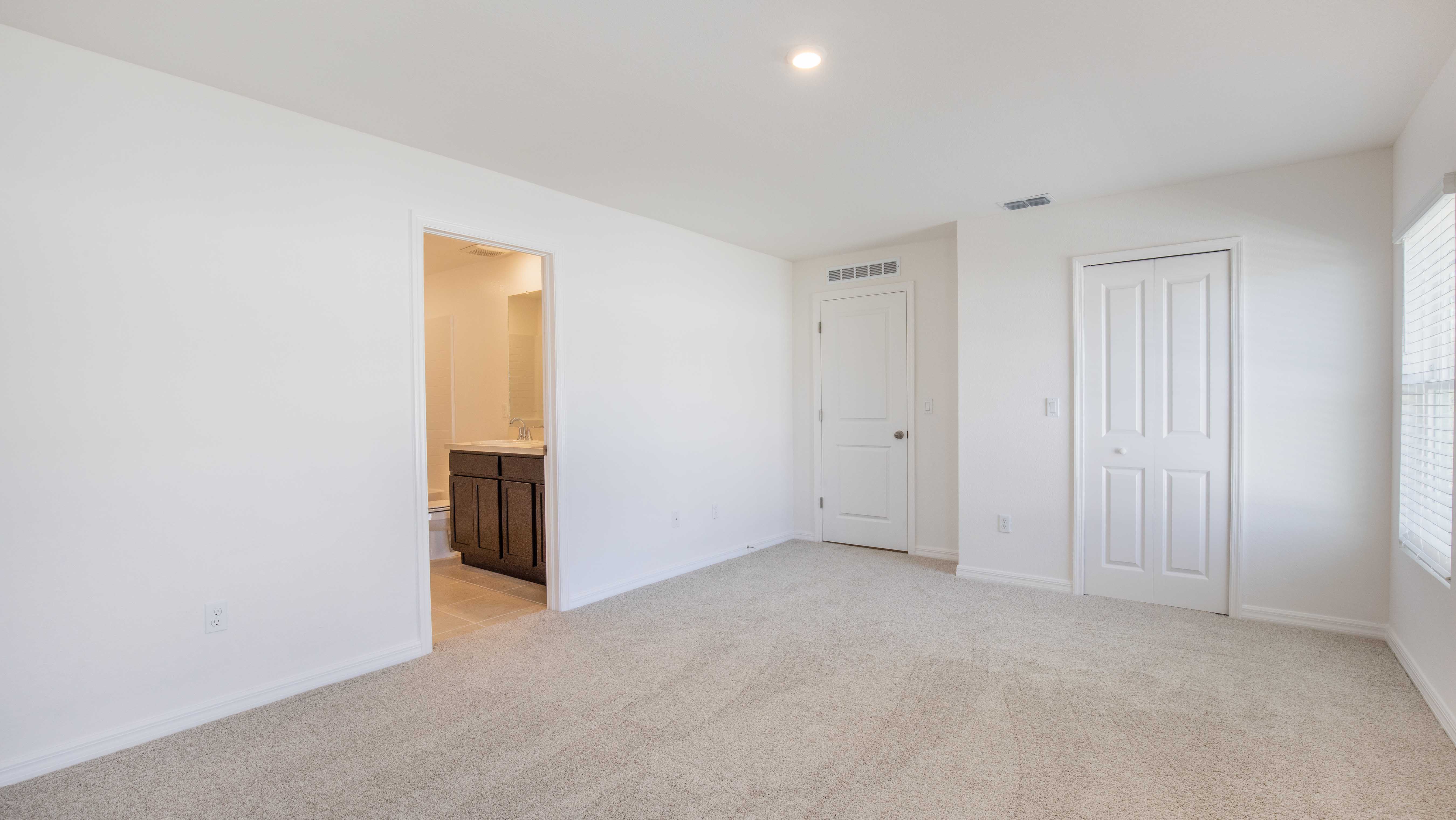 Empty, freshly painted bedroom with beige carpet, an adjoining bathroom, and a closet with white double doors.