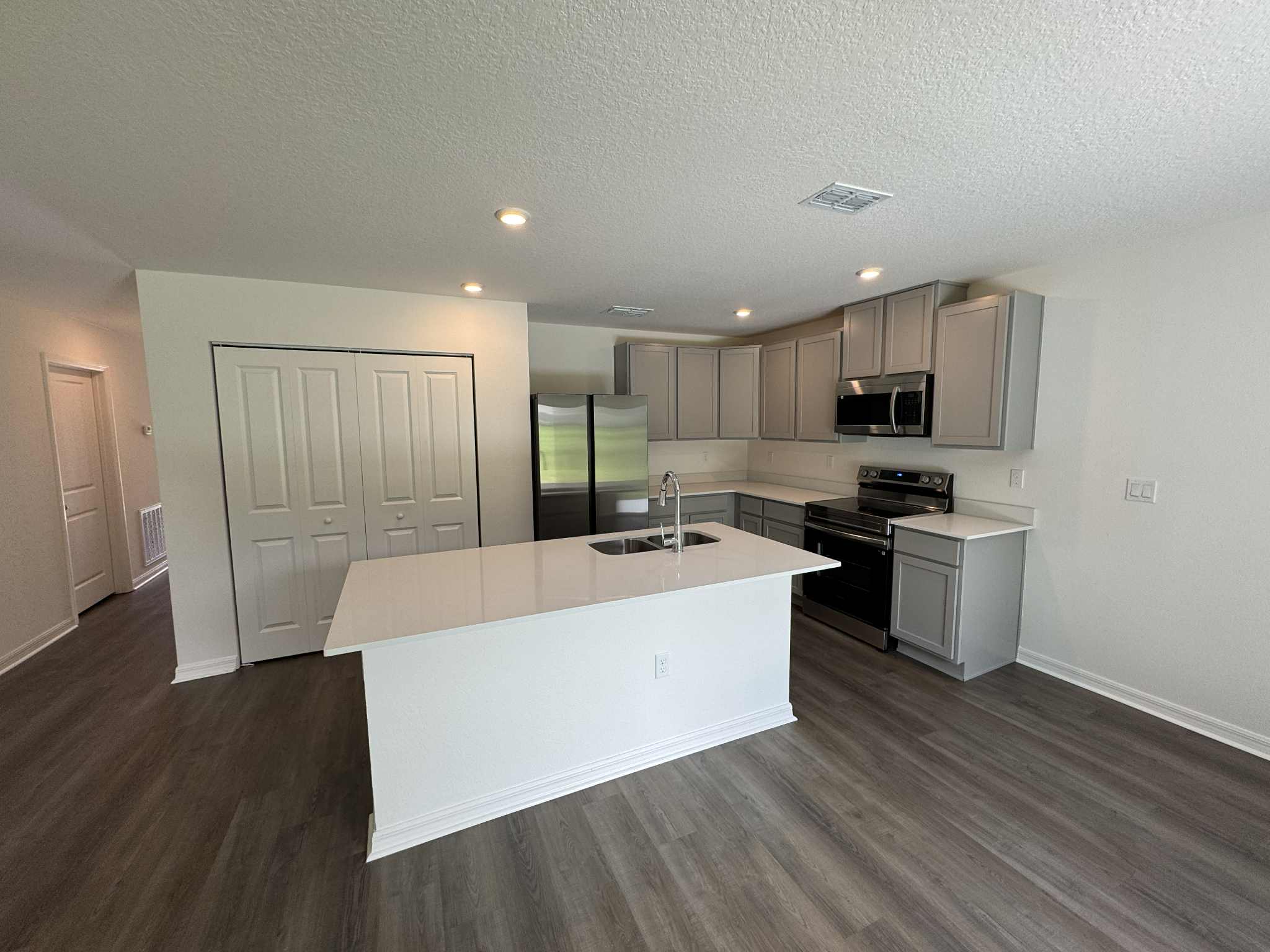 Modern kitchen with gray cabinets, stainless steel appliances, and a central island with a sink, featuring hardwood flooring and recessed lighting.