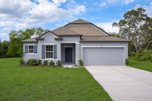 A modern single-story home with a two-car garage, neatly landscaped yard, and gray exterior, set against a backdrop of lush greenery and a cloudy blue sky.