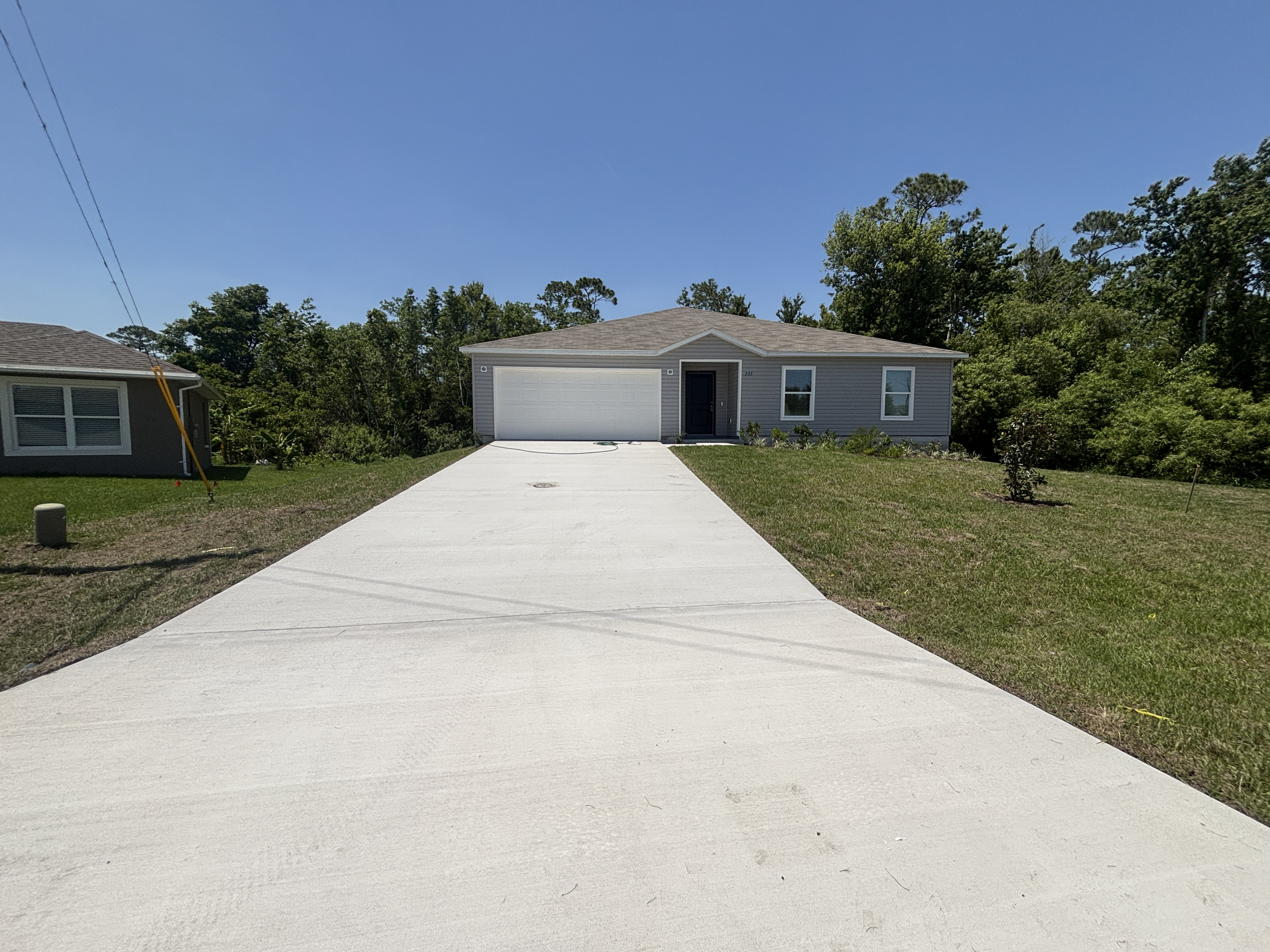Single-story gray house with a wide driveway, surrounded by green grass and trees under a clear blue sky.