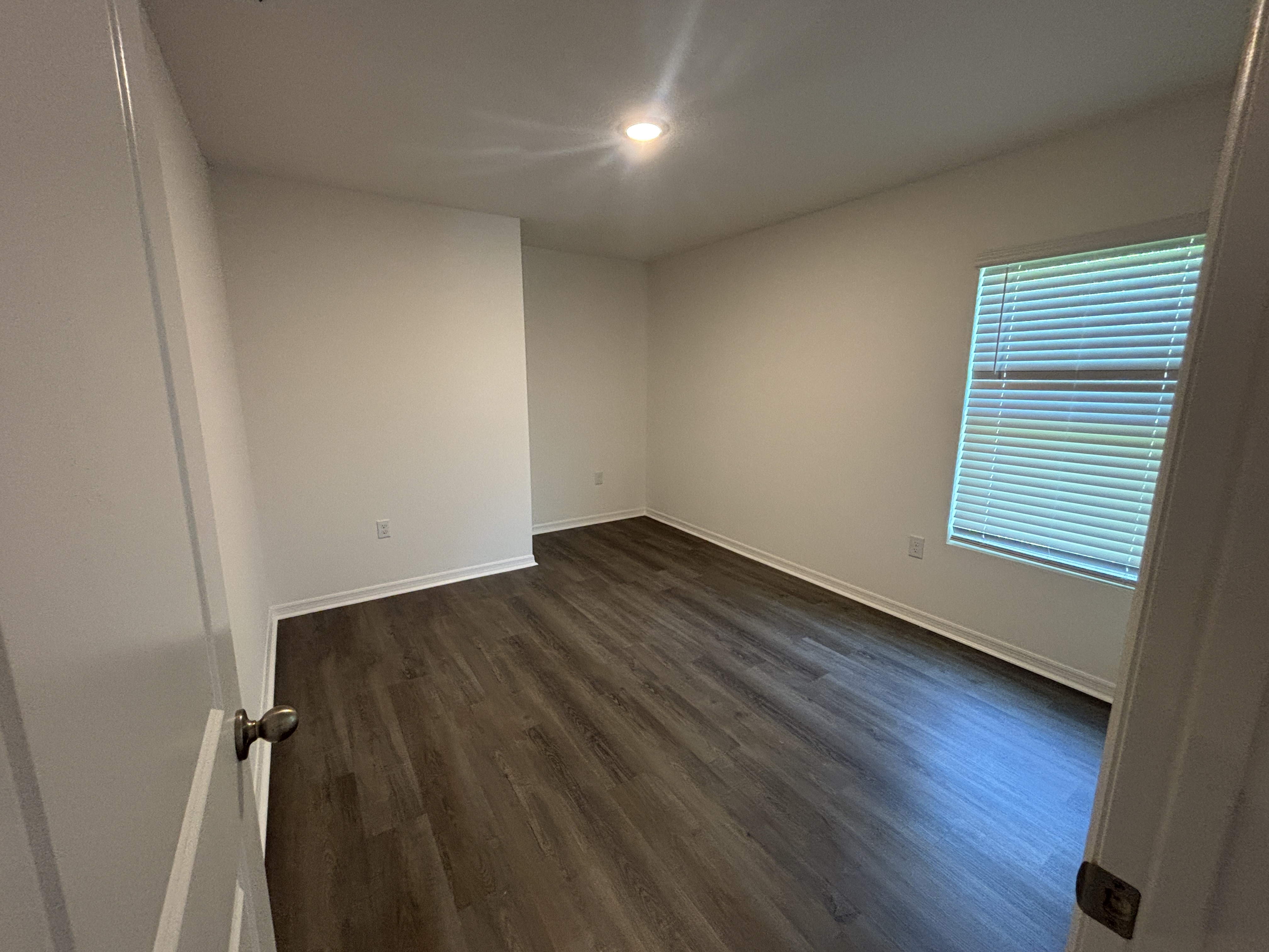 Empty room with white walls, dark wood flooring, a single ceiling light, and a window with blinds.