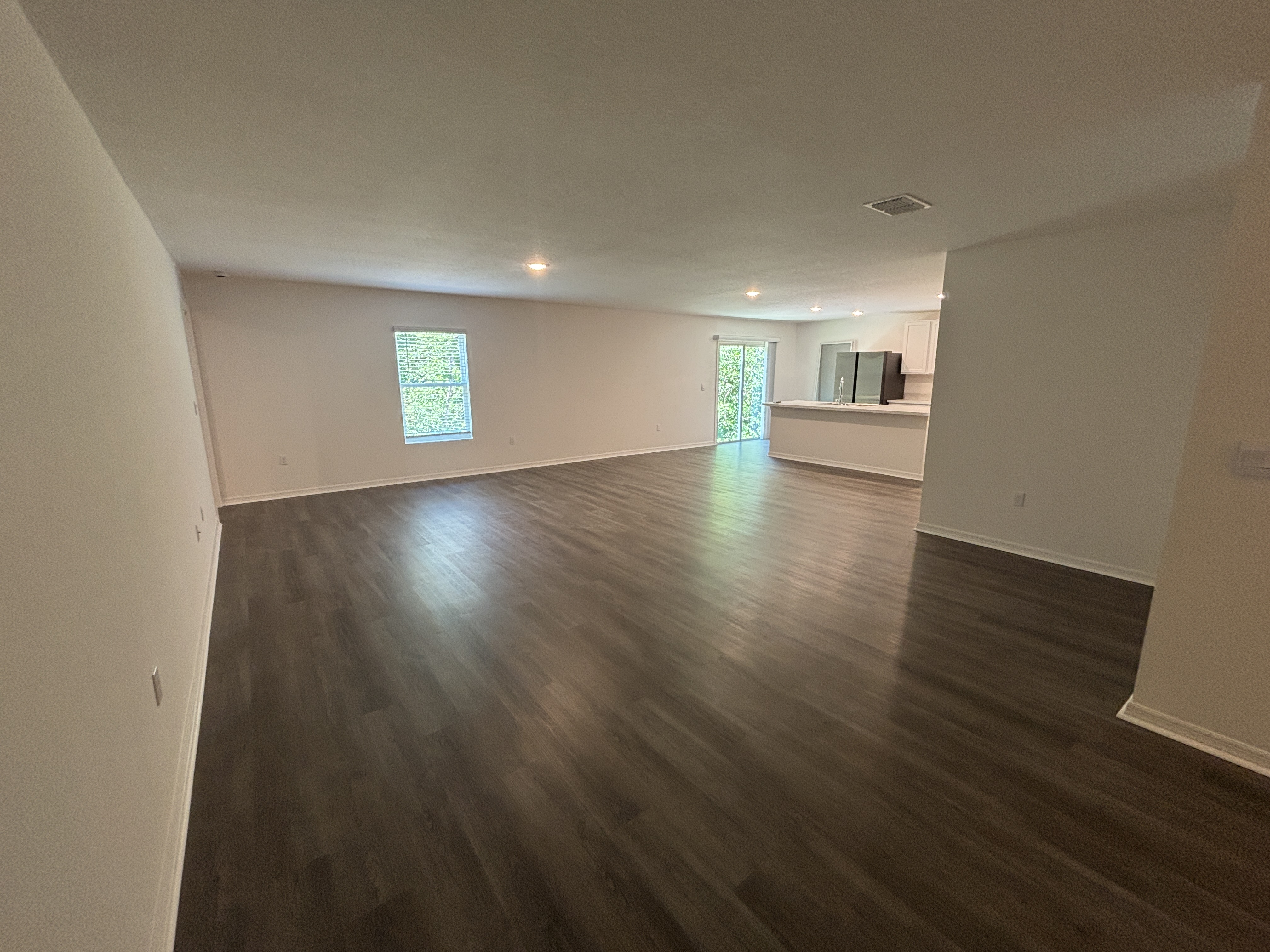 Spacious unfurnished living room with dark wood flooring, white walls, and an open kitchen area featuring modern appliances.